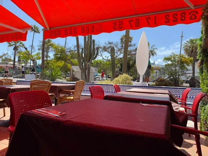 Outdoor restaurant patio with empty tables covered in red tablecloths, red and wicker chairs, an orange awning, and desert landscaping with cacti, palm trees, and clear blue sky in the background.