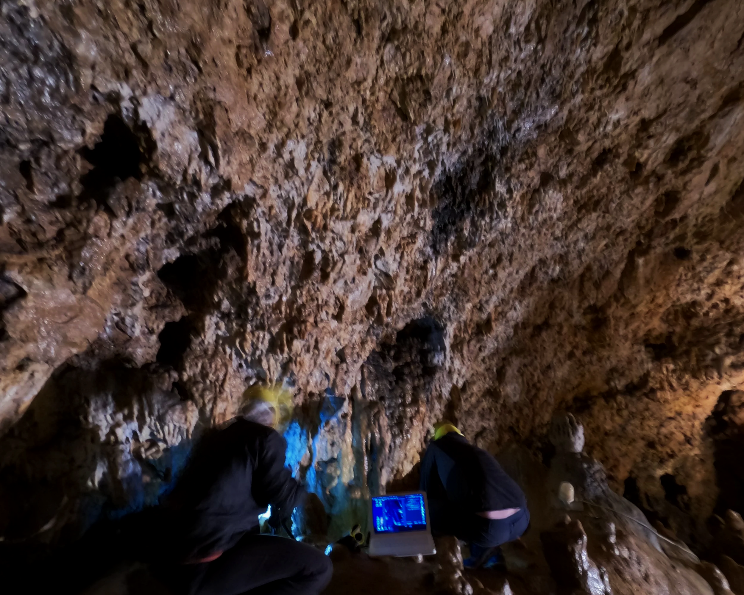 Blick in eine Höhle mit unebenen, nassen Wänden, an denen Wasser tropft. Am Boden sind Steine und Erde. Es sind Personenschuhe sichtbar, sowie eine Person, die einen Laptop mit einer blauen Anzeige benutzt.