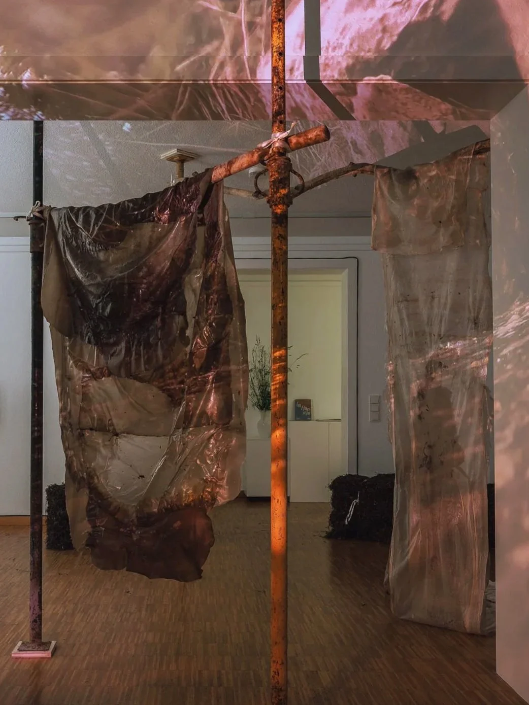 Kunstinstallation mit rustikalen Metallstangen, durchsichtigen Stoffbahnen mit abstrakten Mustern, Projektionslicht an der Decke und einem inneren Raum mit Wanddeko im Hintergrund.