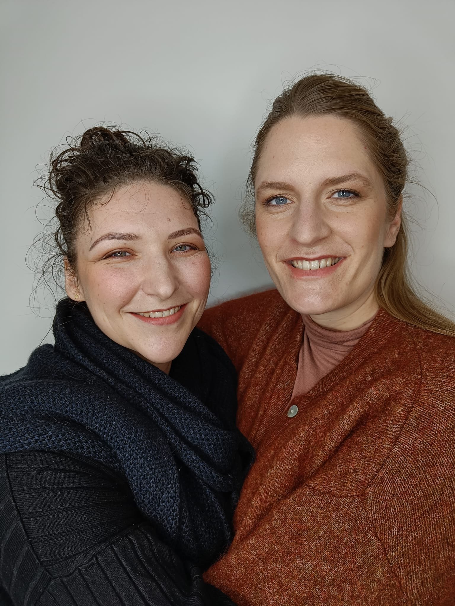 Two people smiling, one wearing a dark scarf and sweater, the other in a rust-colored cardigan.