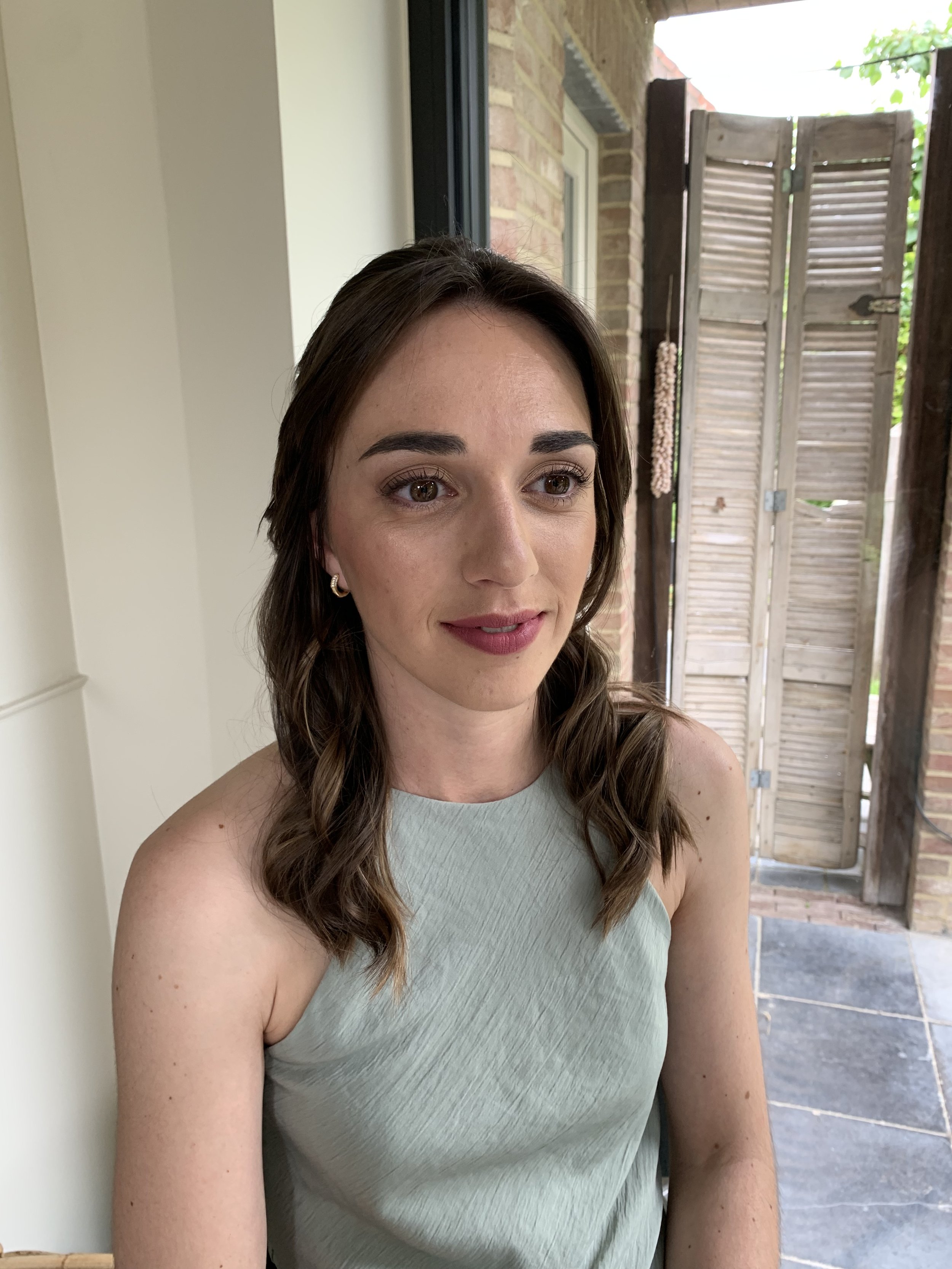 A woman with brown hair wearing a sleeveless gray dress is sitting indoors near a window. She has a neutral expression. In the background, there is a wooden shutter and a brick wall.