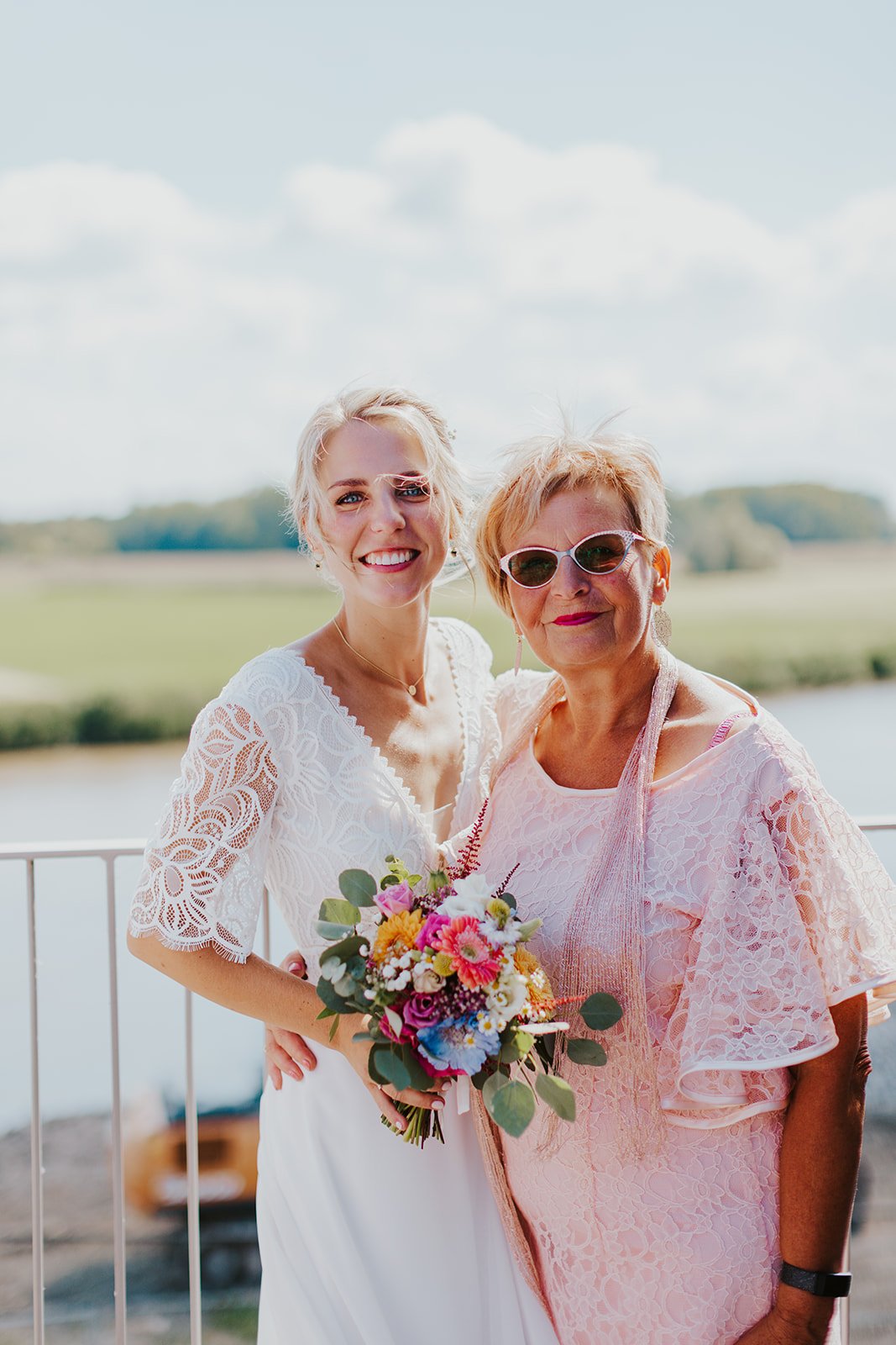 Bride in white dress holding bouquet with woman in pink lace dress outdoors, smiling.