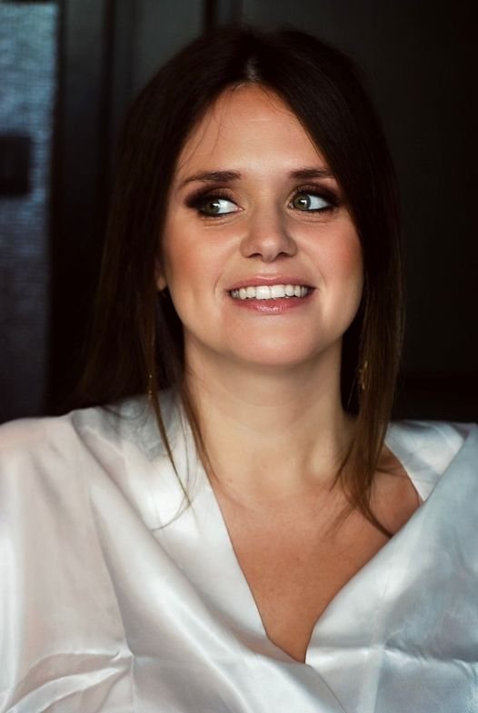 Woman with long brown hair smiling, wearing a white blouse, in an indoor setting.