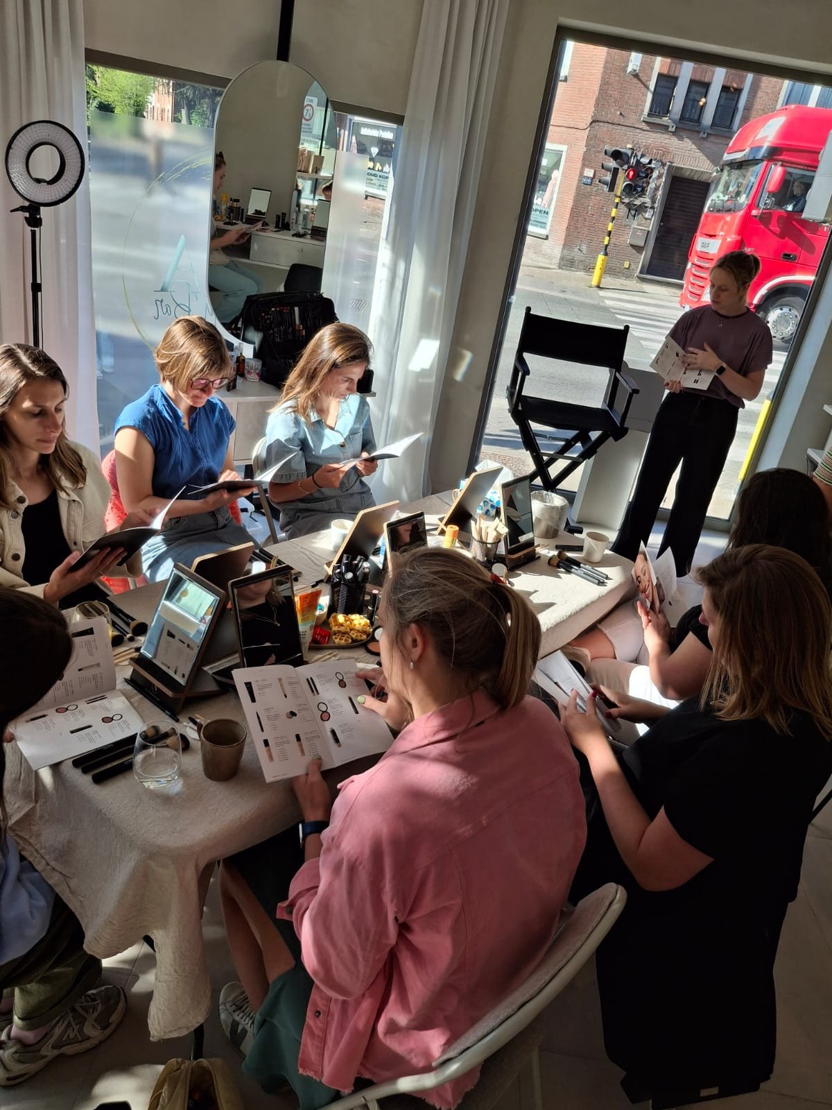 A group of people sitting around a table in a well-lit room with large windows. They are looking at documents and using tablets. A person is standing and presenting next to a director's chair. Ring light and various items like cups and snacks are on