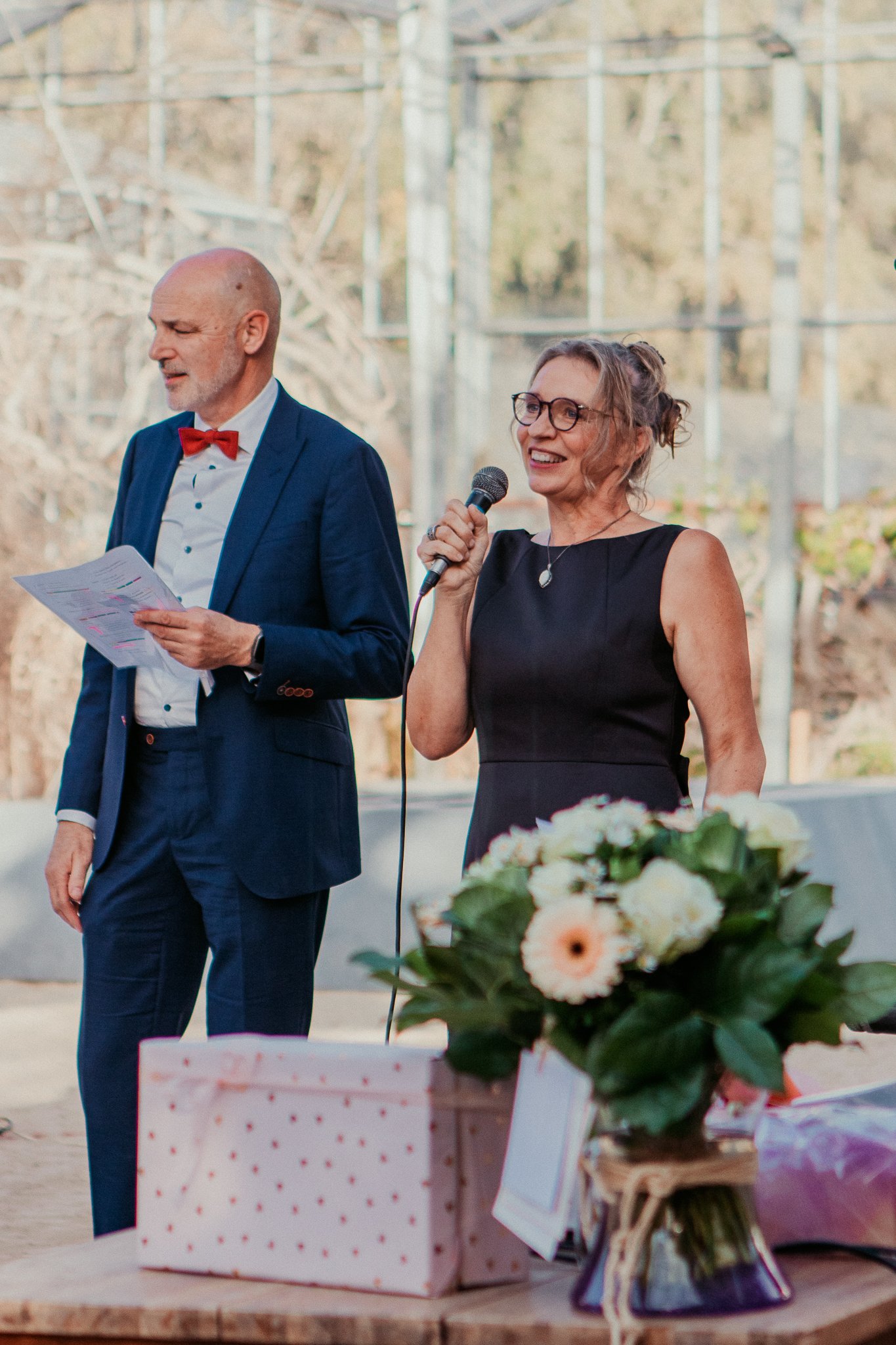 Two people standing beside a table with flowers and a gift box, speaking into a microphone at an outdoor event.
