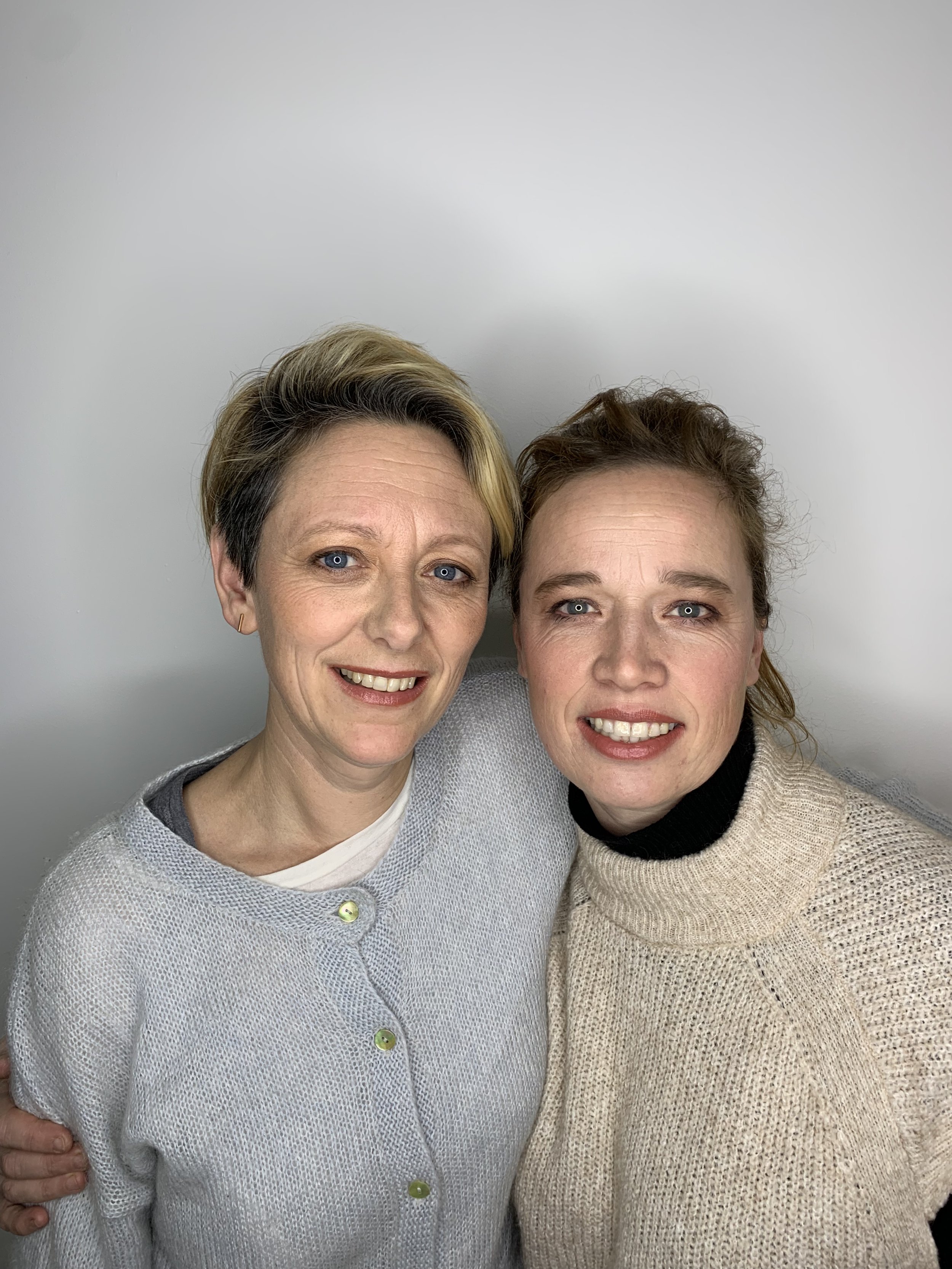 Two smiling people wearing knitted sweaters, posing together against a plain background.