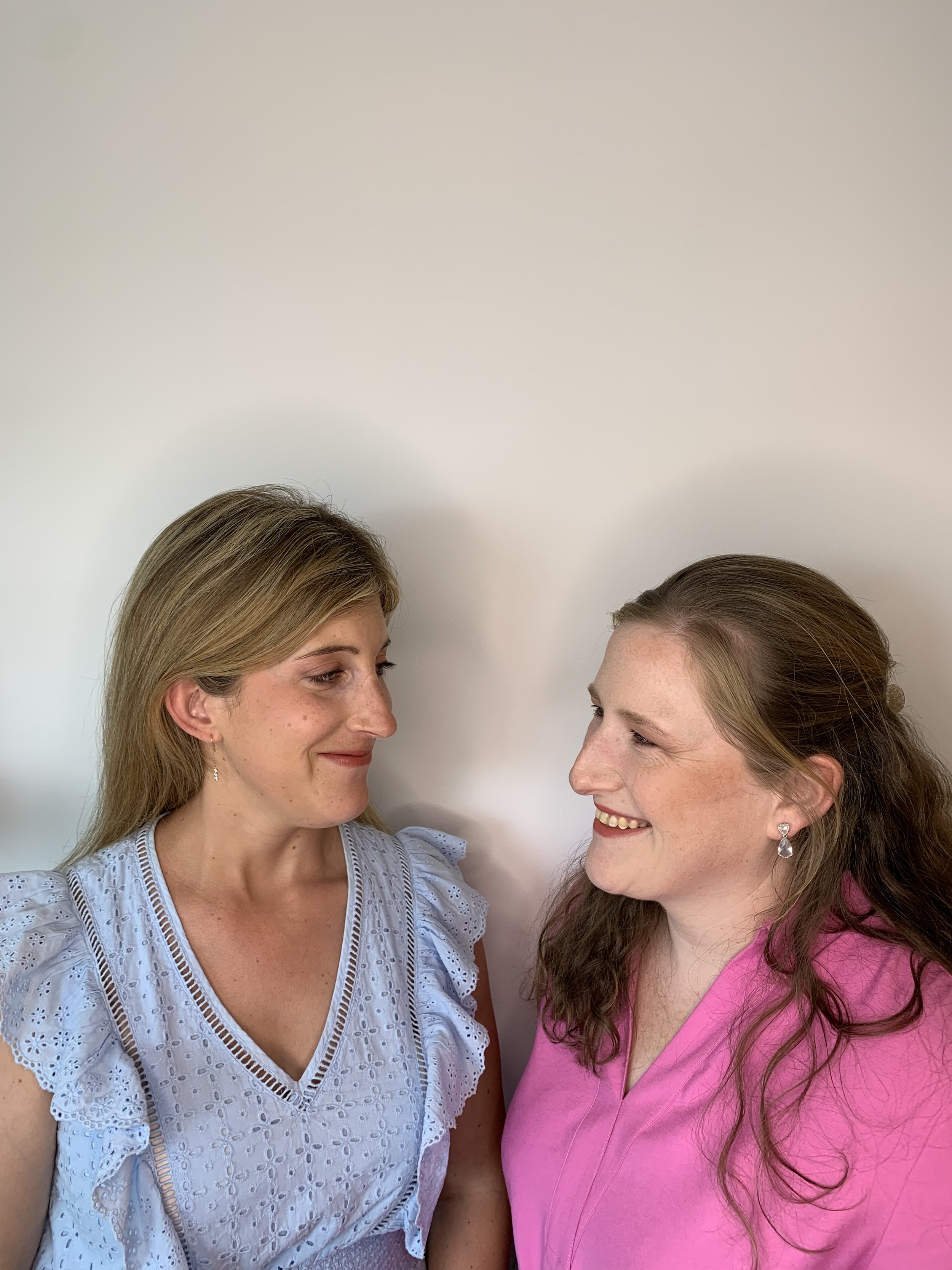 Two women smiling at each other, one in a light blue blouse, the other in a pink shirt, against a plain background.