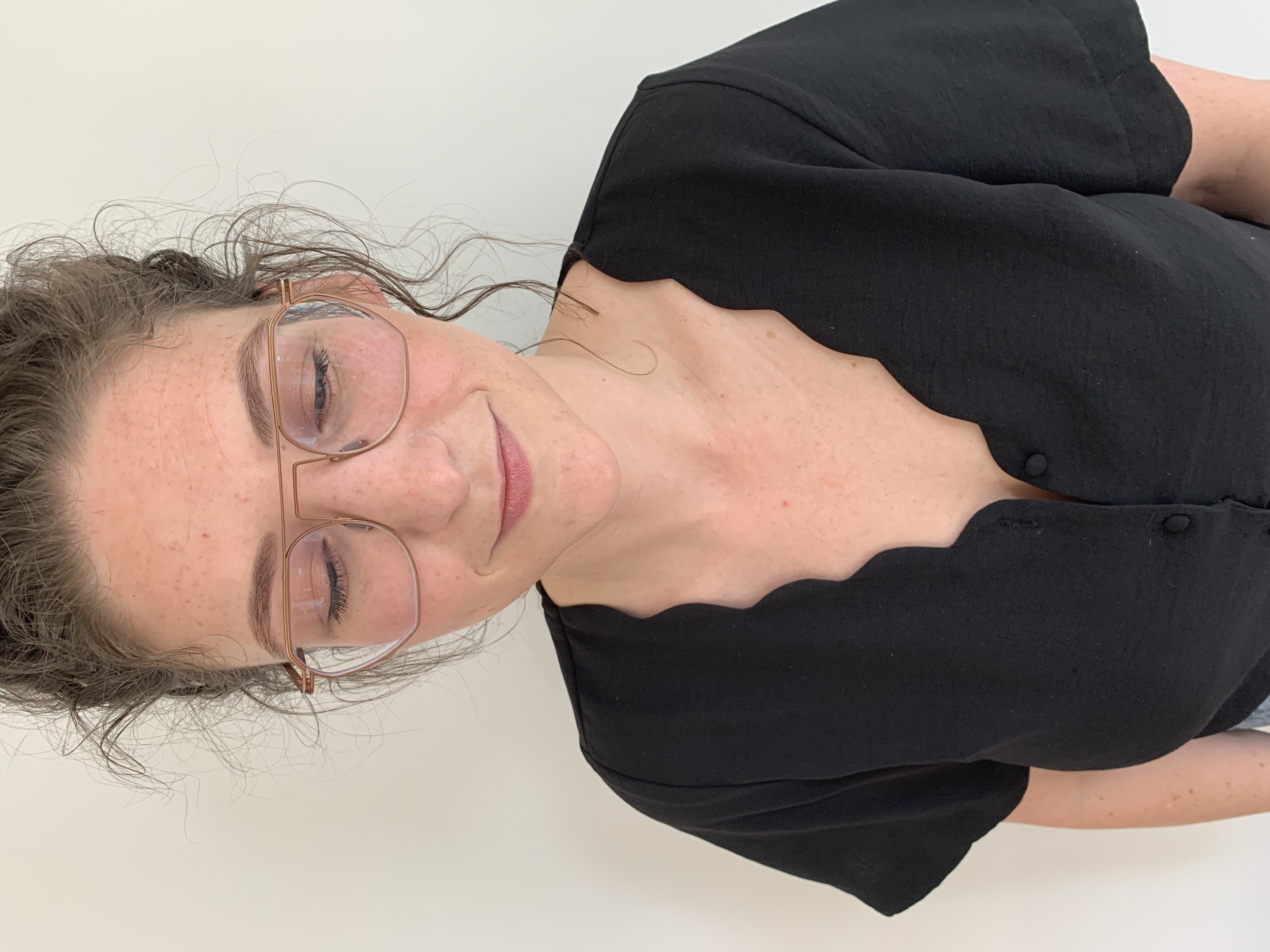 Woman with glasses and wavy hair wearing a black dress with a scalloped neckline, looking down against a plain background.