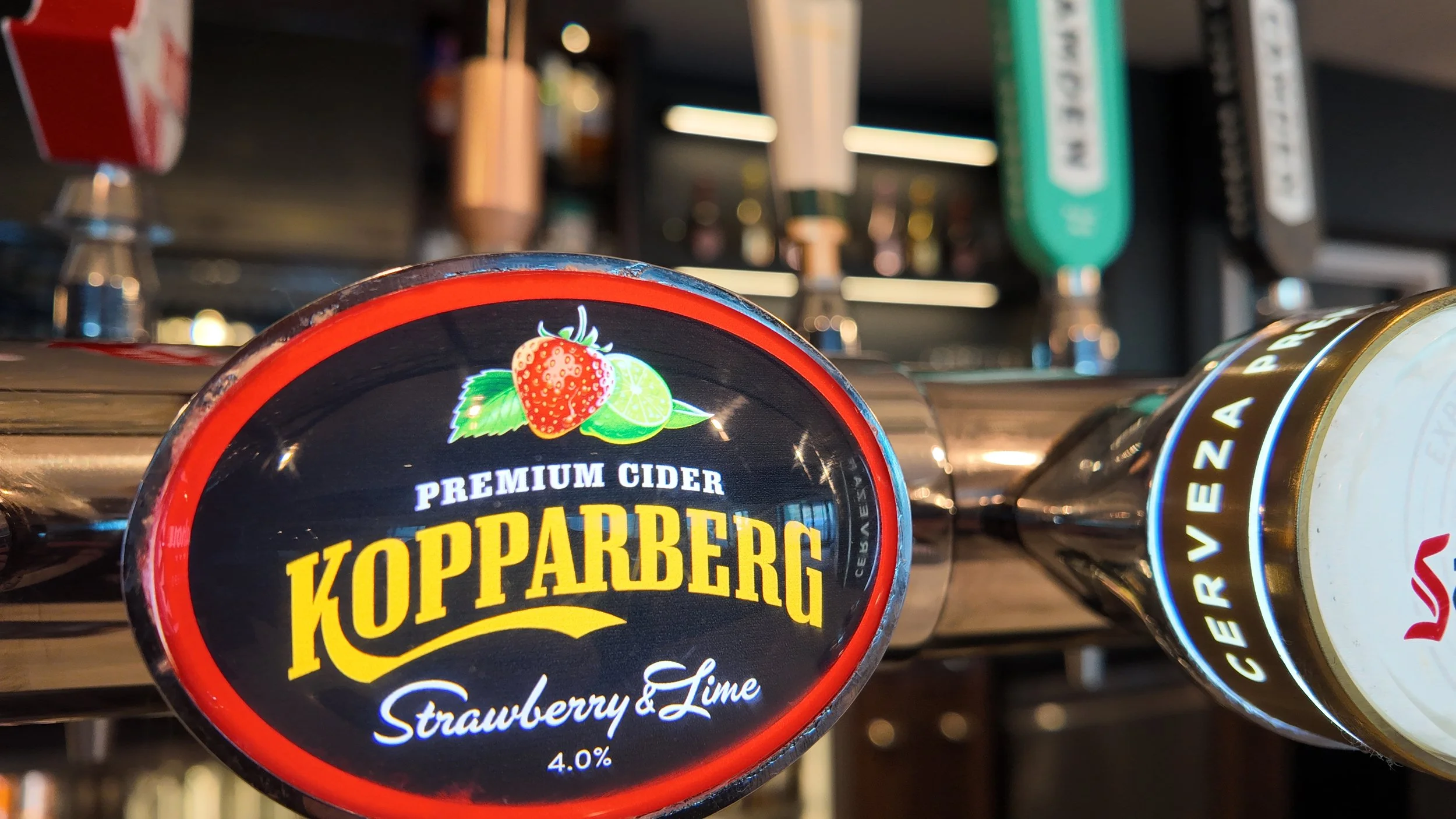 Close-up of beer taps, highlighting a Koppaberg strawberry & lime premium cider tap with a strawberry, lime, and green leaves illustration on the label. Other taps are in the background.