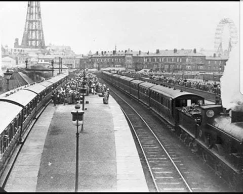 Black and white photo of a busy train station in Paris with a steam locomotive, train cars, people on the platform, the Eiffel Tower, and a Ferris wheel in the background.