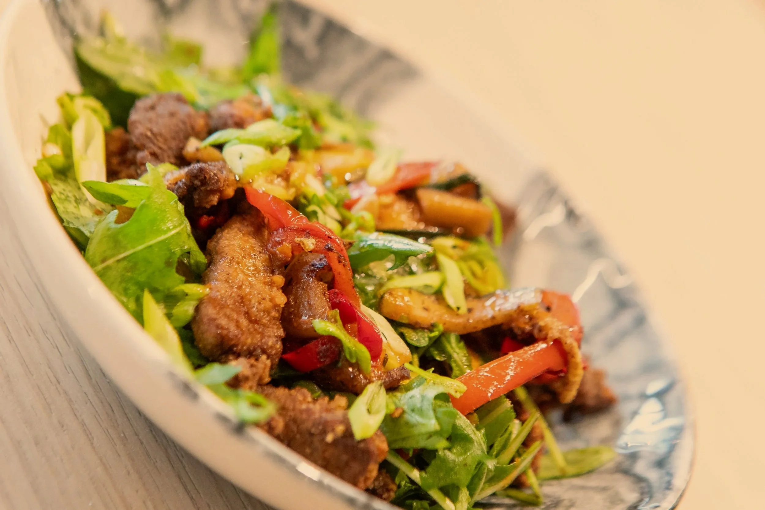 A close-up of a colorful Asian beef stir-fry with vegetables, including green onions, red bell peppers, and broccoli, served in a white oval dish.