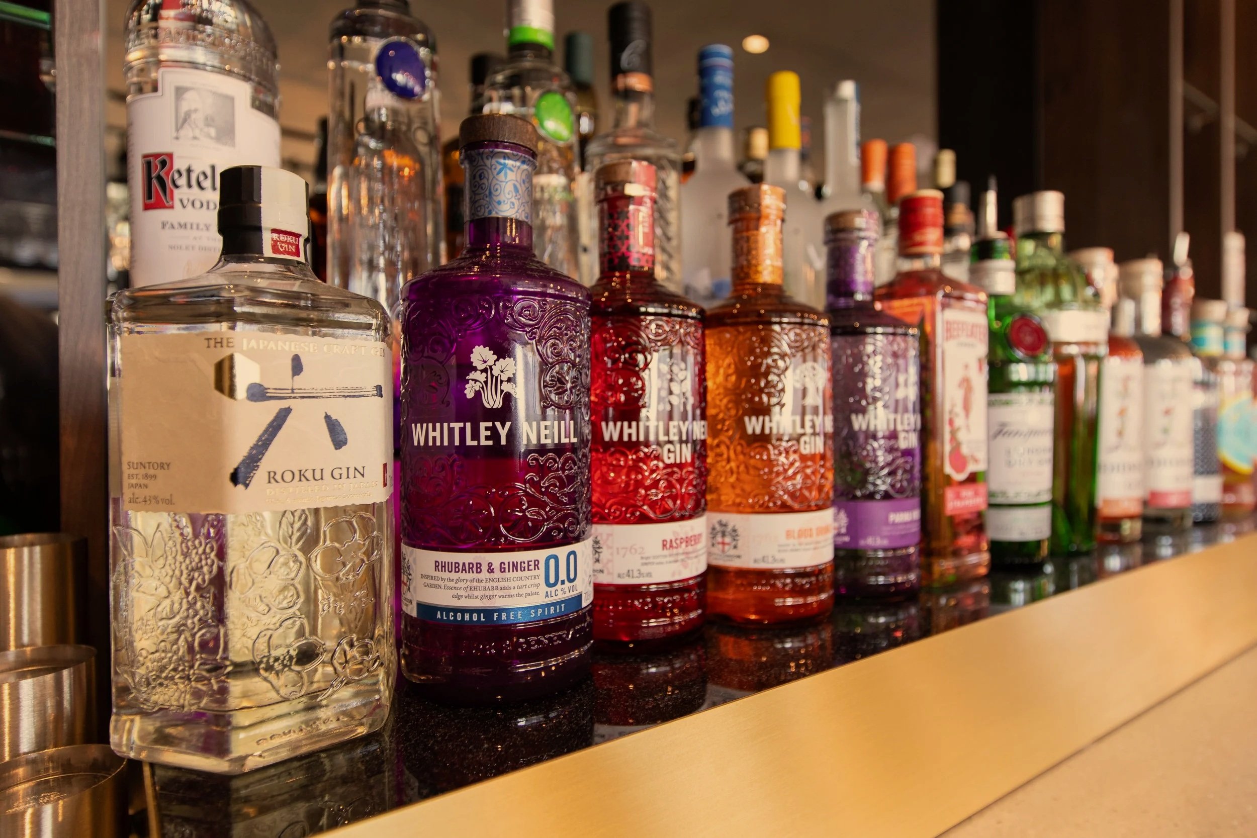 A line of colorful liquor bottles on a bar counter, including Whitley Neill gin and Roku gin, with various other bottles in the background.