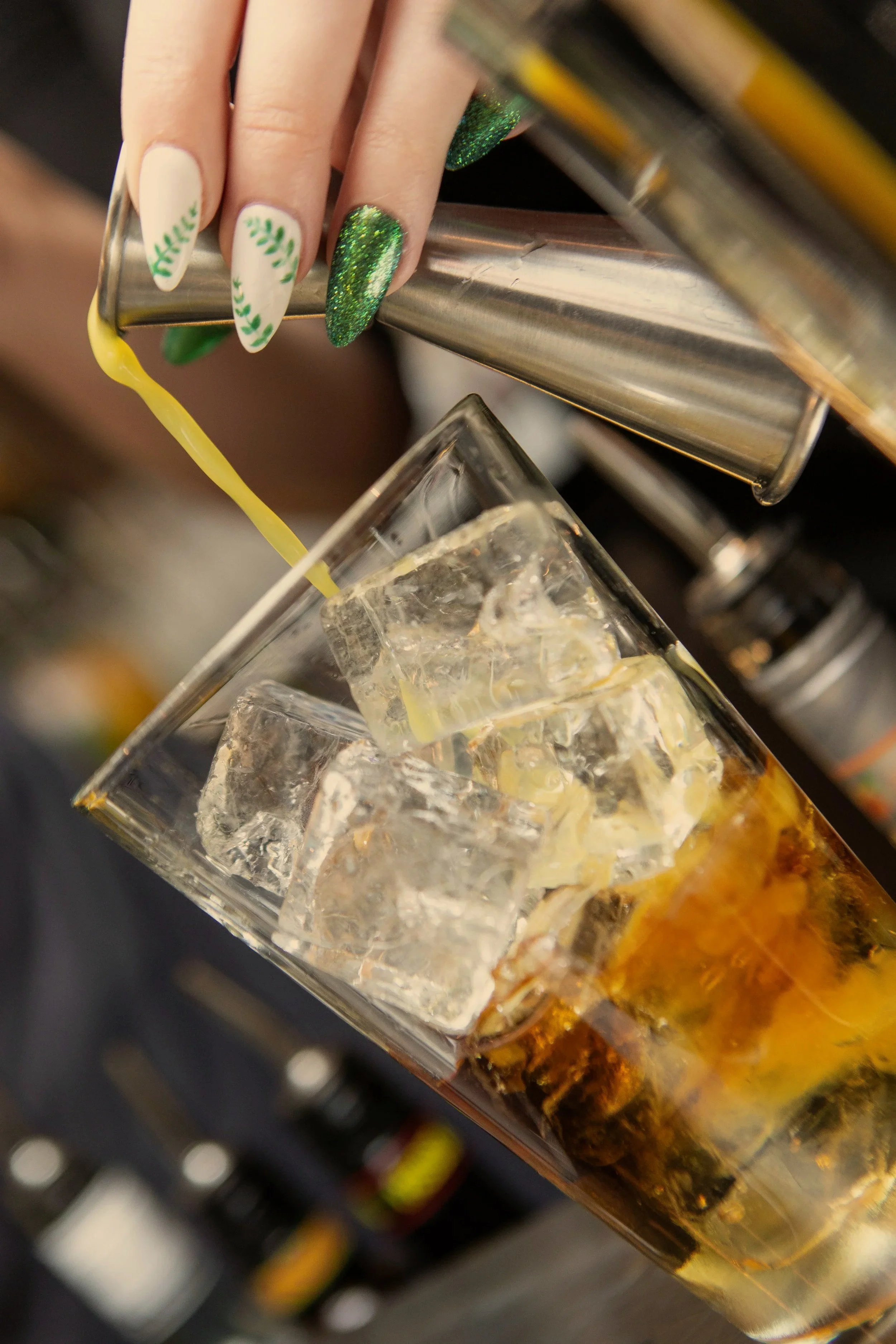 A bartender pours a yellow beverage into a glass with ice cubes, using a metal cocktail shaker.