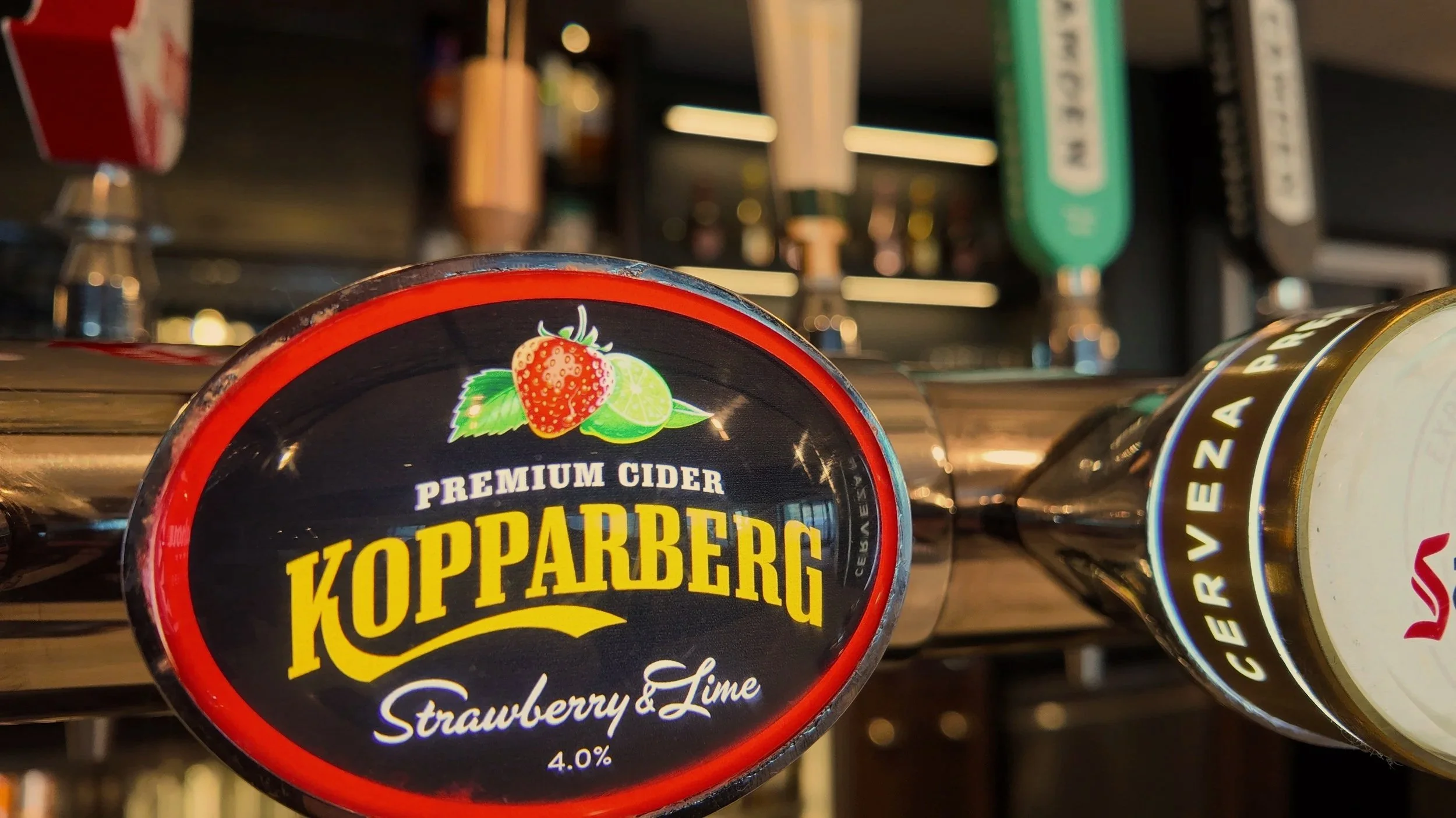 Close-up of beer taps, highlighting a Koppaberg strawberry & lime premium cider tap with a strawberry, lime, and green leaves illustration on the label. Other taps are in the background.