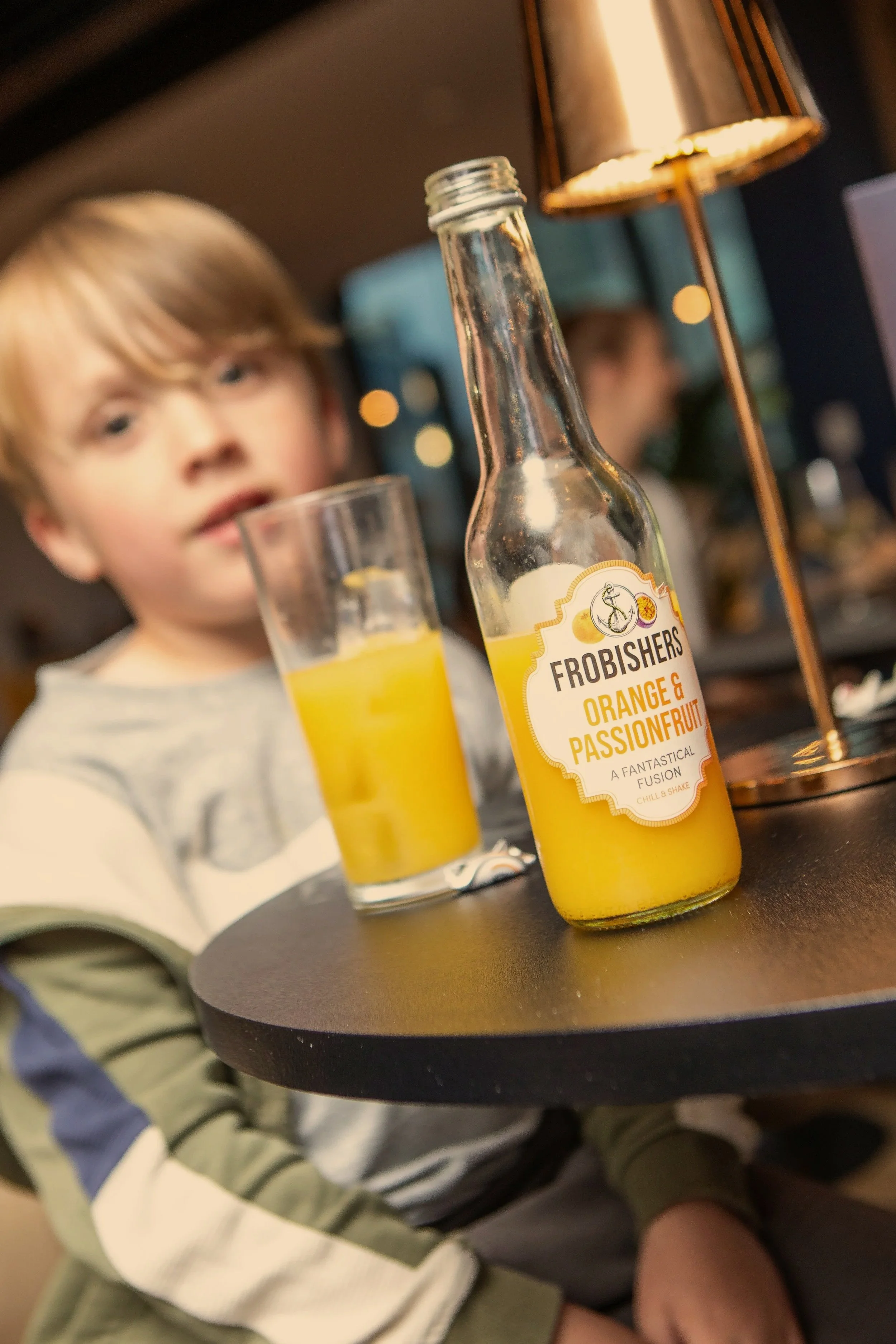 A young boy in a gray and green sweatshirt sitting at a black table with a glass of orange juice and a bottle labeled "FROBISHERS ORANGE & PASSIONFRUIT, A FANTASTICAL FUSION, CHILL & SHAKE," in a restaurant or cafe setting.