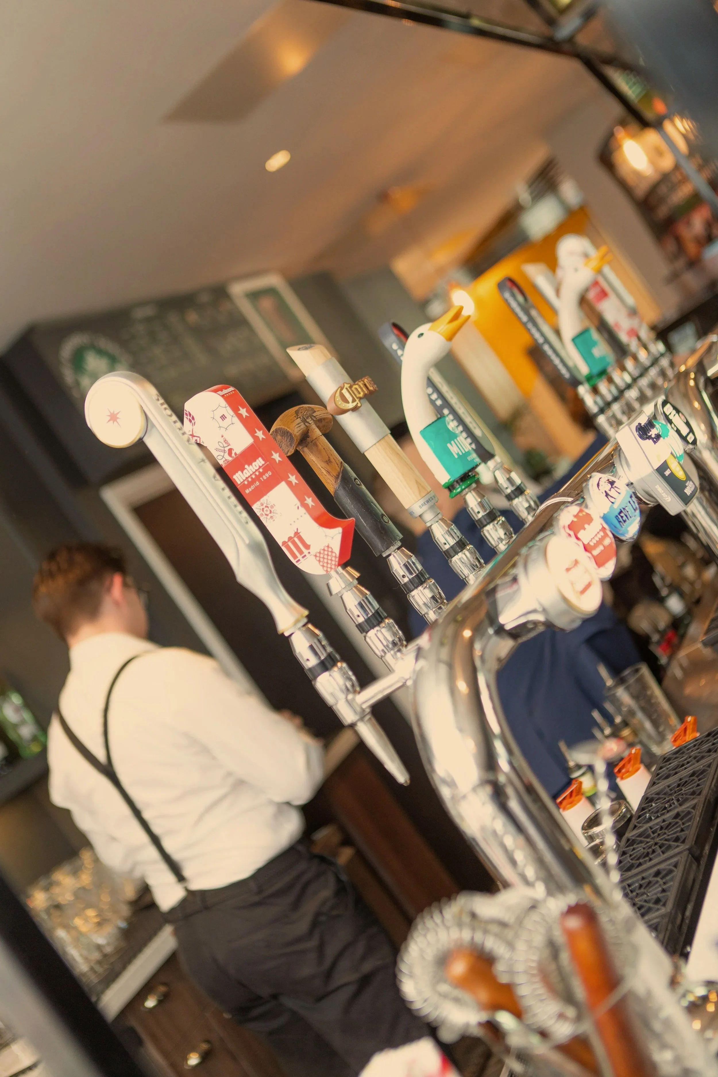 Multiple beer taps on a bar, with various decorative handles, in a restaurant or bar setting. A bartender in a white shirt and black suspenders is visible in the background.