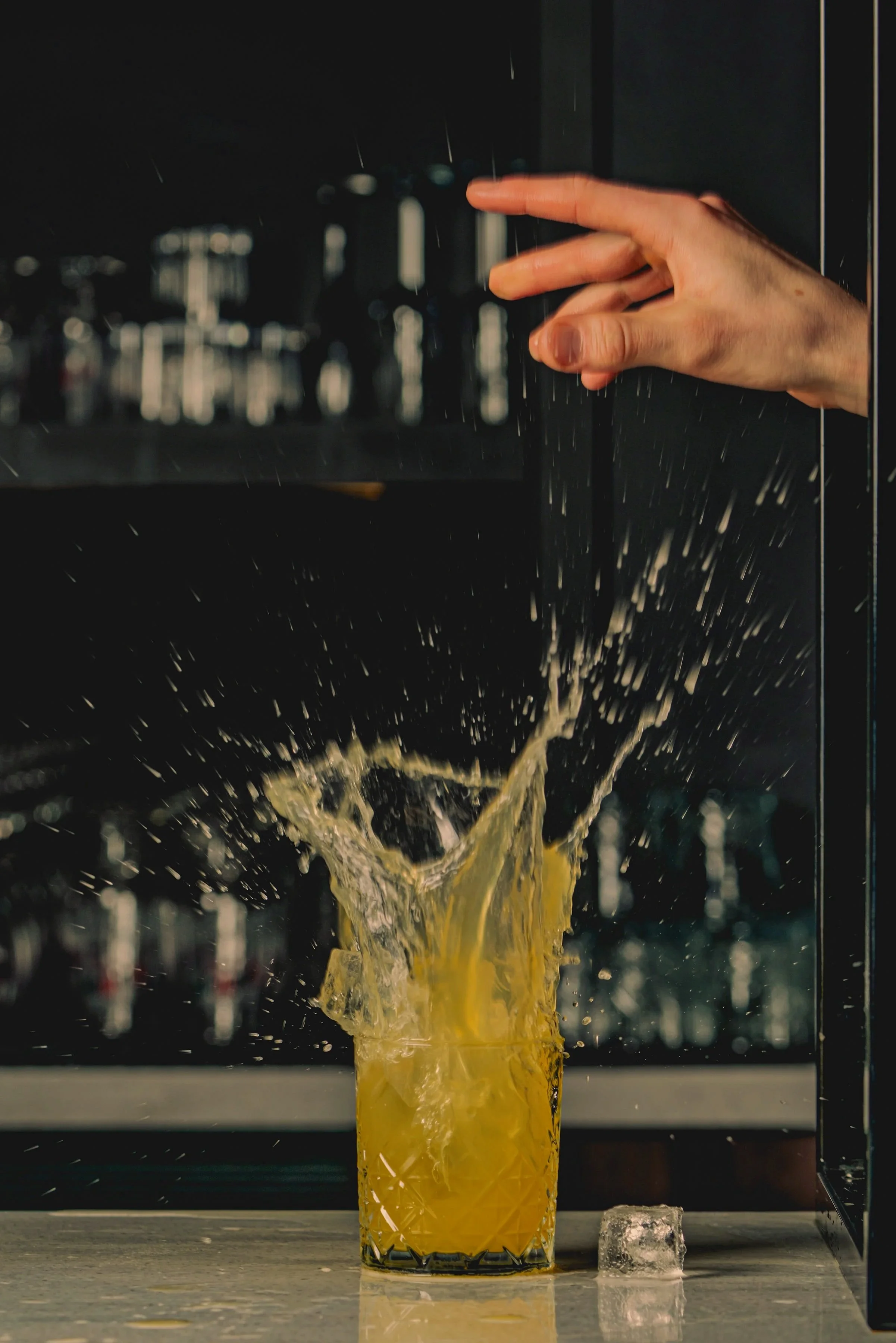 A hand is seen dropping ice into a glass of orange juice, causing a splash.