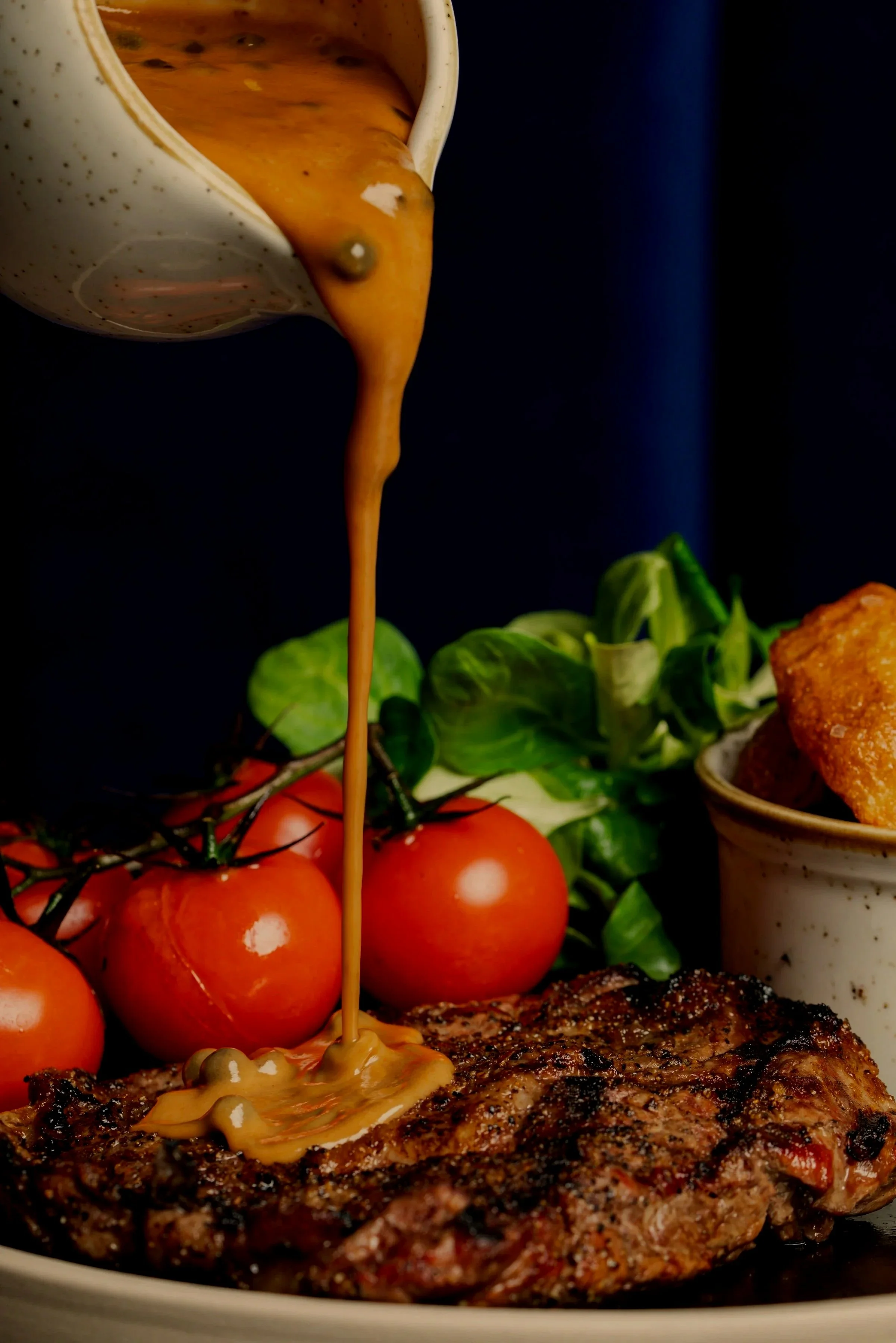 Grilled steak with gravy being poured over it, accompanied by cherry tomatoes, green leafy vegetables, and a bowl of chips.