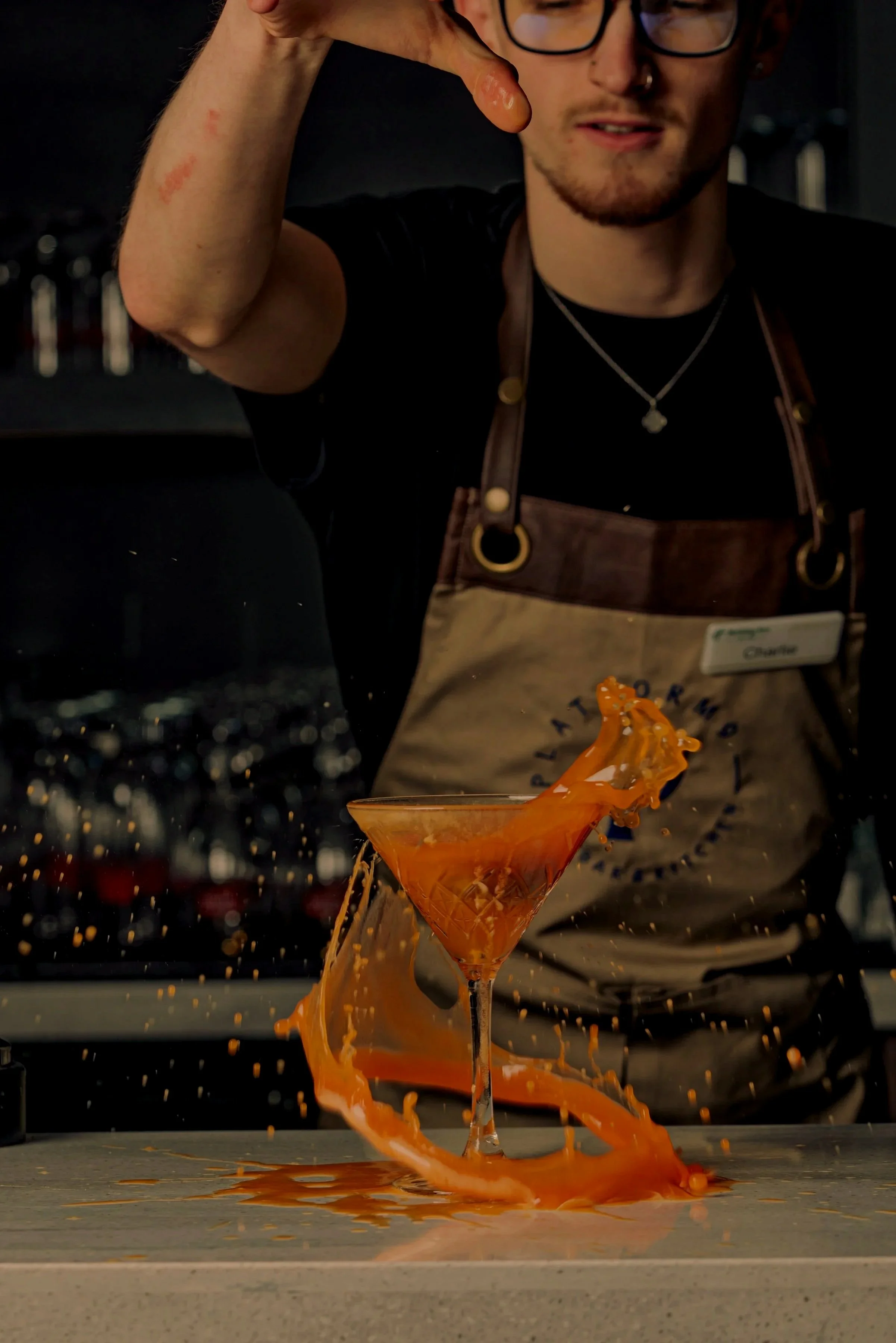 A bartender wearing glasses and a brown apron is pouring orange liquid into a martini glass, causing a splash that spills over the glass and onto the countertop.