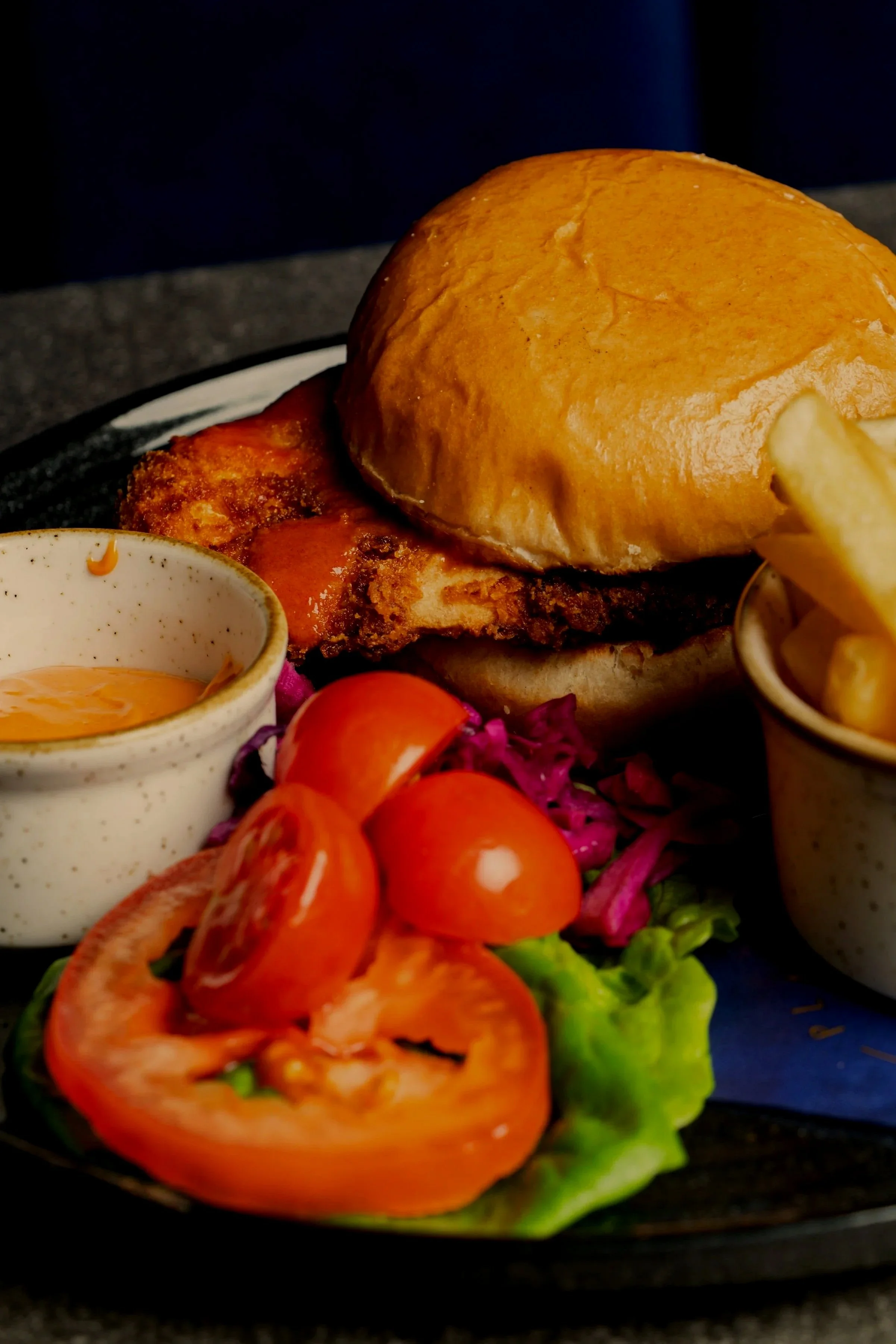 A cheeseburger with a bun, lettuce, tomato, fries, and side condiments on a black plate.