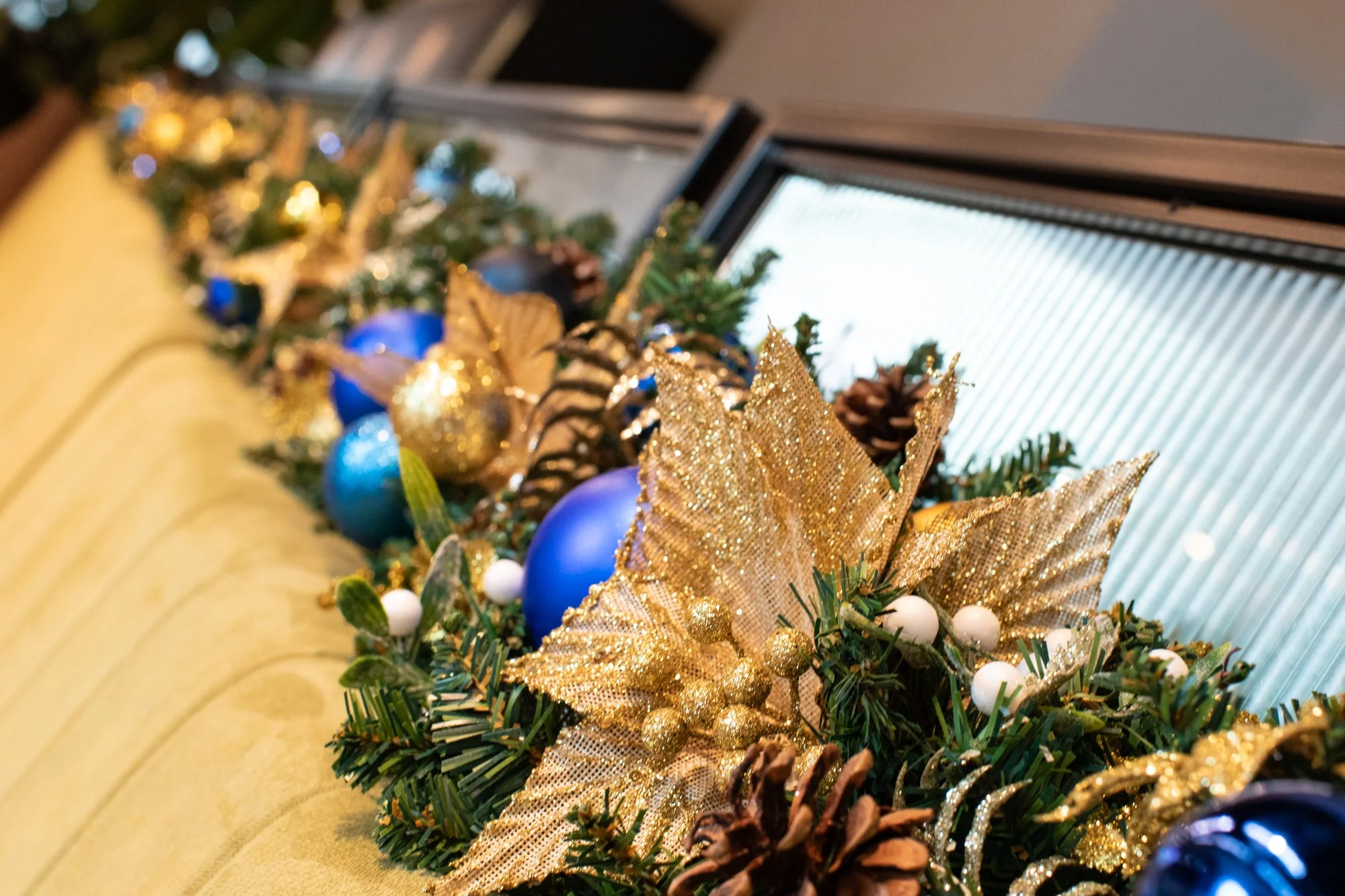 Close-up of a decorated Christmas wreath with gold ribbons, pinecones, white berries, and blue ornaments near a window.