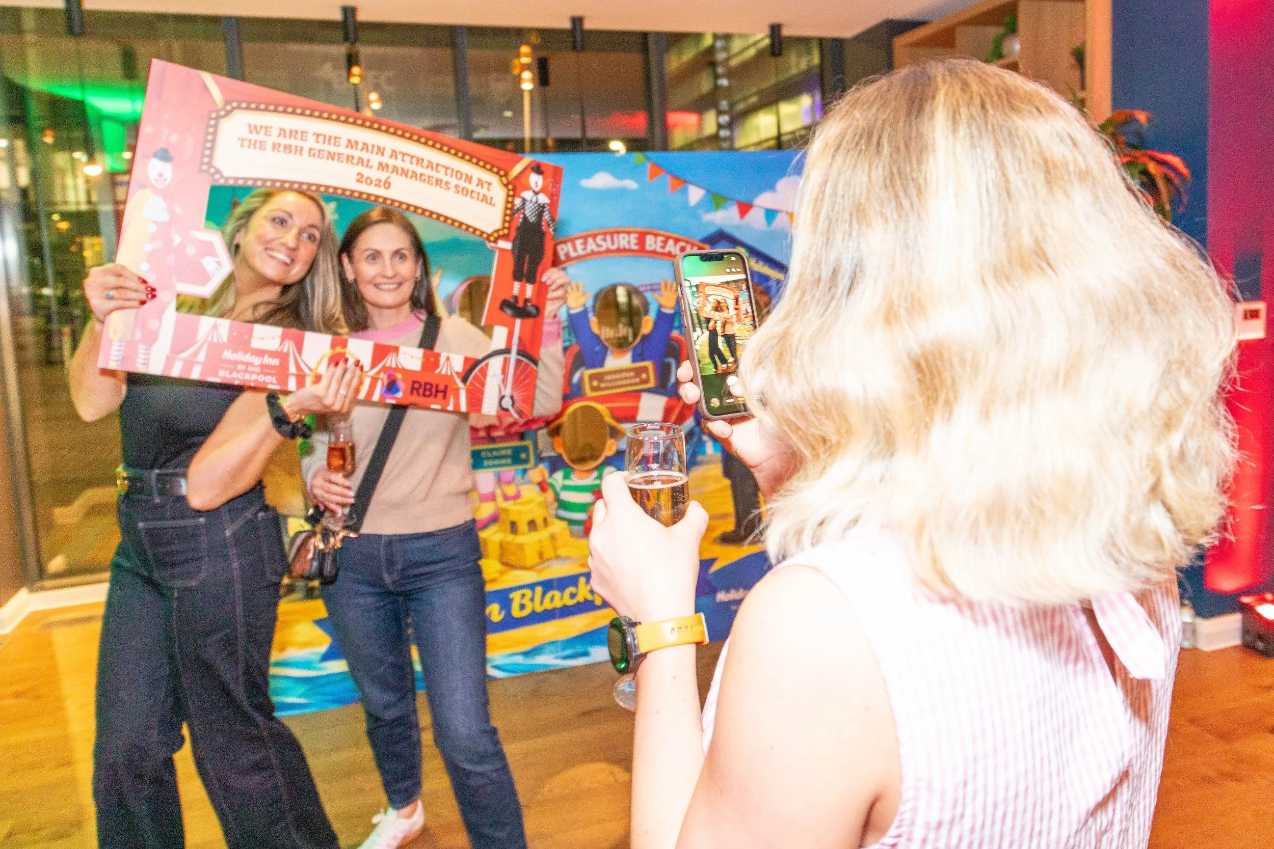 Two women are posing with a festive photo frame at an indoor event, with a third person taking their picture. The background features colorful decor and a play area for children.