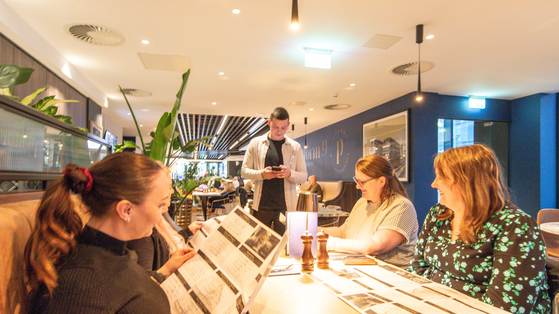 People sitting at a restaurant table, looking at menus, while a man stands behind them using his phone inside a modern, well-lit restaurant.