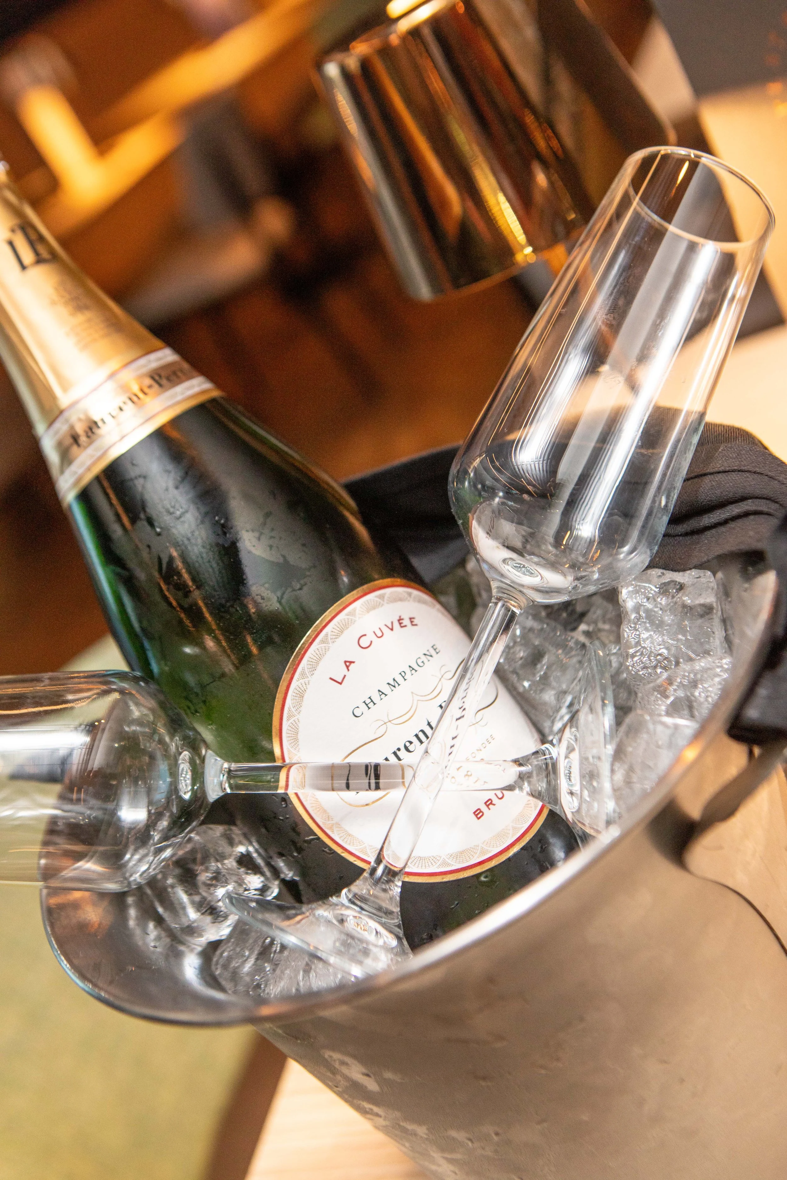A bottle of champagne in a metal ice bucket filled with ice, with two empty champagne glasses placed nearby, all on a wooden surface.