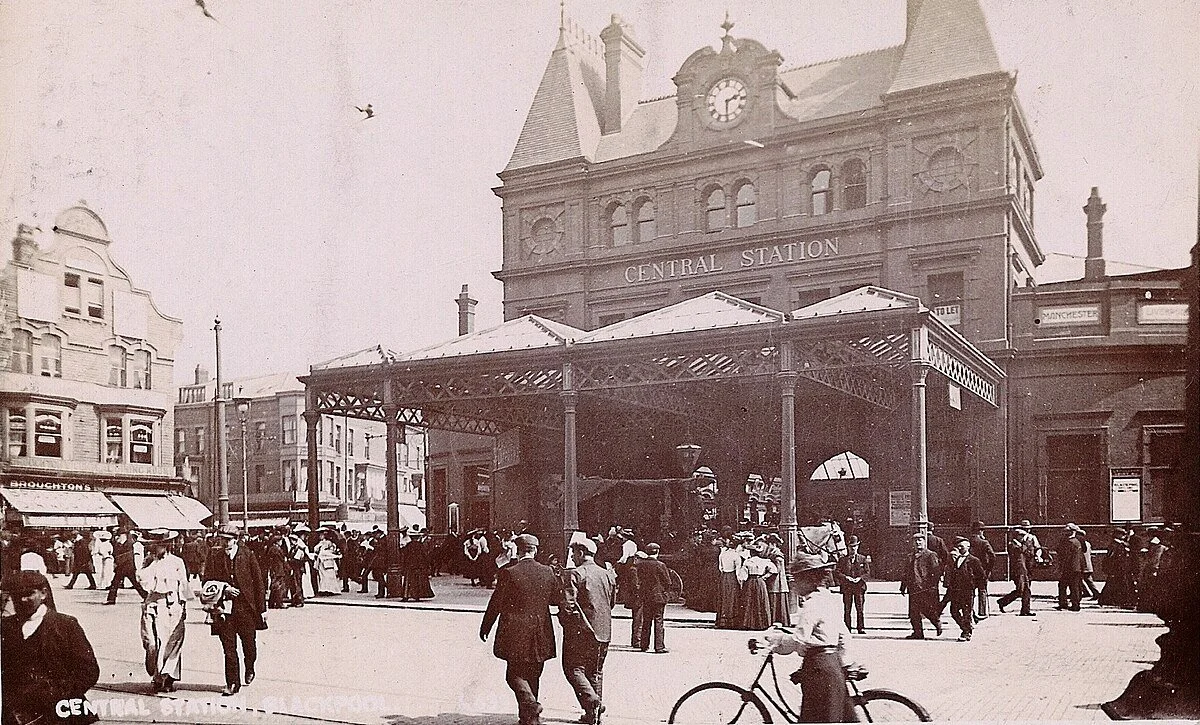 Historical black and white photo of a busy city street outside a train station labeled 'Central Station'. The station building has a clock and a small tower. There is a covered entrance and numerous people walking, standing, and riding bicycles in front of the station, with shops and buildings on either side.