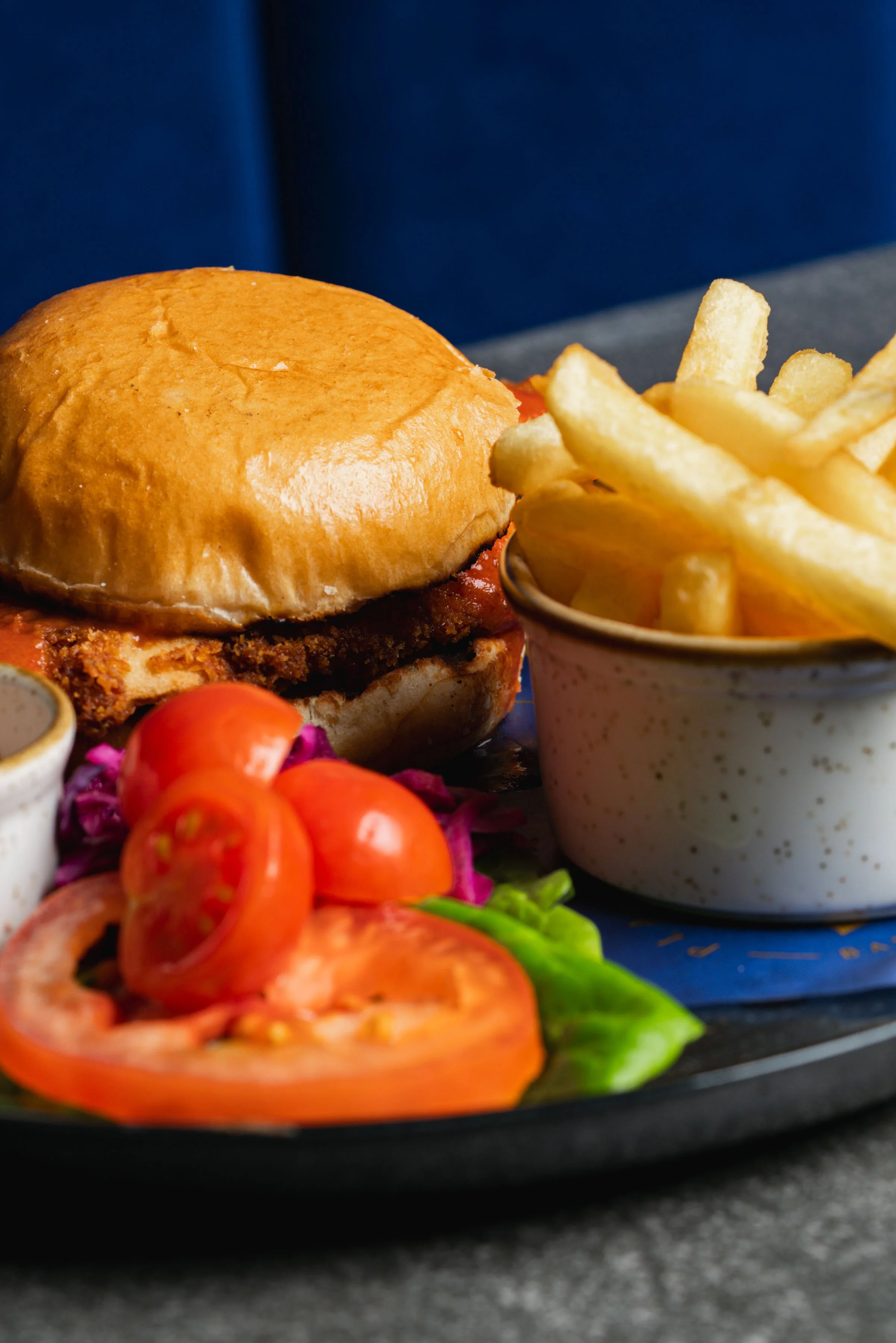 Close-up of a burger with melted cheese, accompanied by French fries and slices of tomato and lettuce on a black plate.