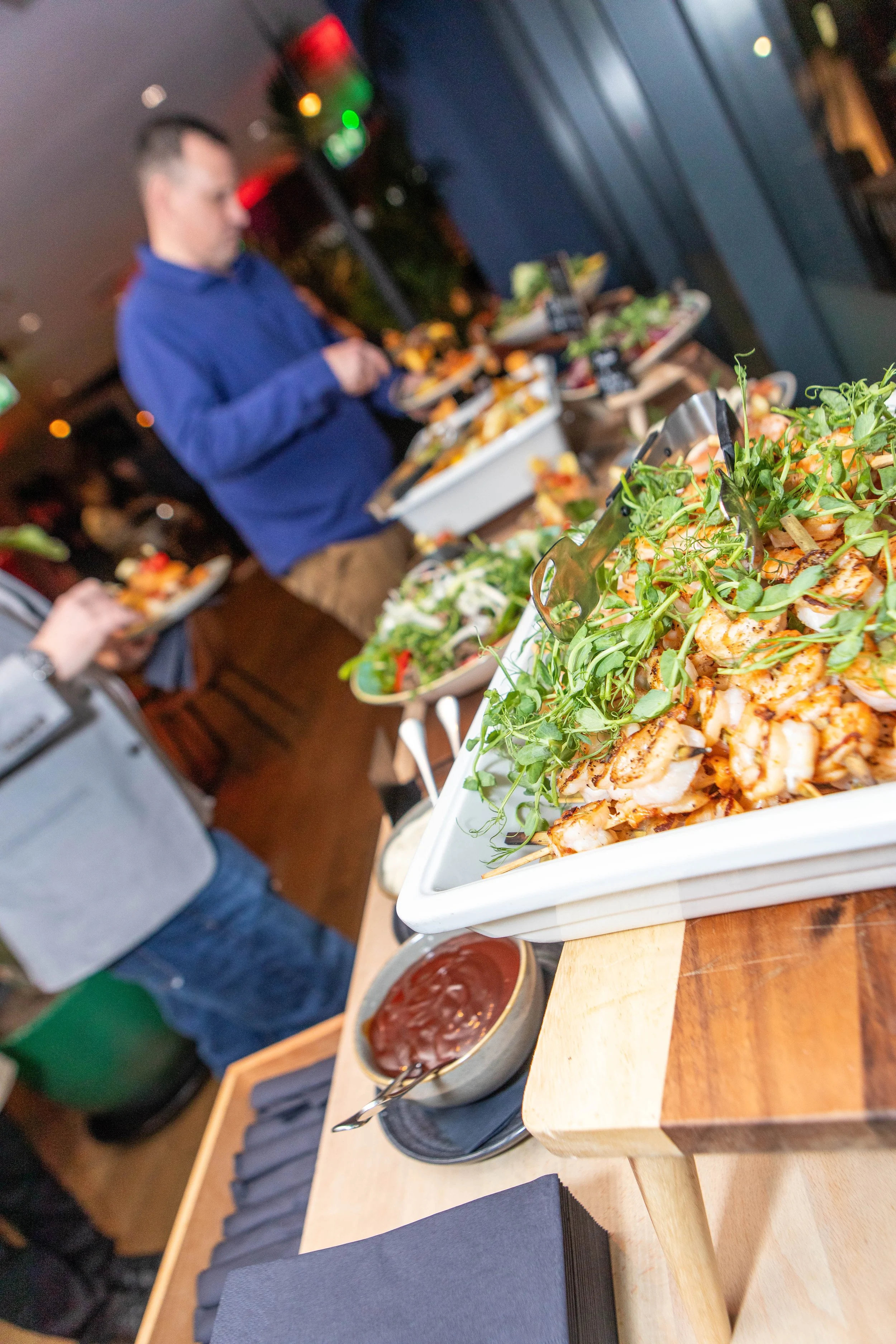 A buffet table with various dishes, including a seafood platter with greens, and a bowl of dipping sauce, with people serving themselves in the background at a dining event.