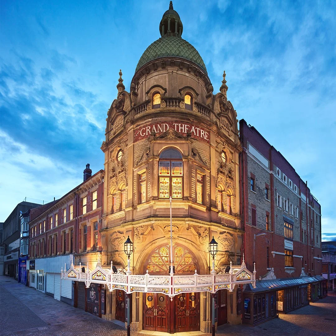 Historic building with a rounded dome labeled 'The Grand Theatre', illuminated at dusk with a decorative white balcony and lamp posts at the entrance.