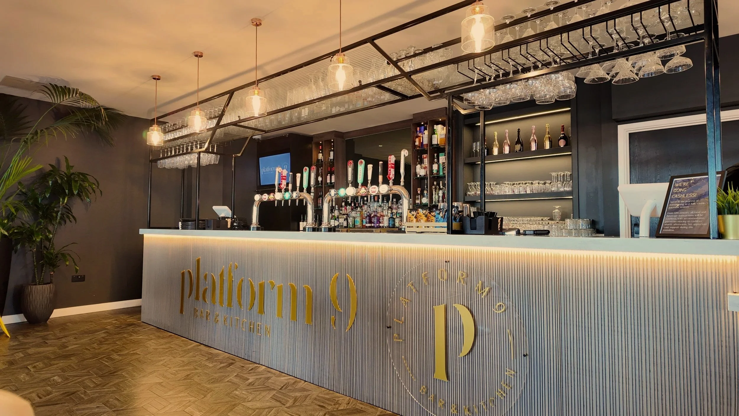 Modern bar with a marble countertop, illuminated from below, displaying the word 'platform (9) BAR & KITCHEN' in gold lettering. Behind the bar are shelves filled with liquor bottles, glassware, and beer taps. The ceiling has hanging glass pendant lights, and there are green potted plants on the left side of the bar.