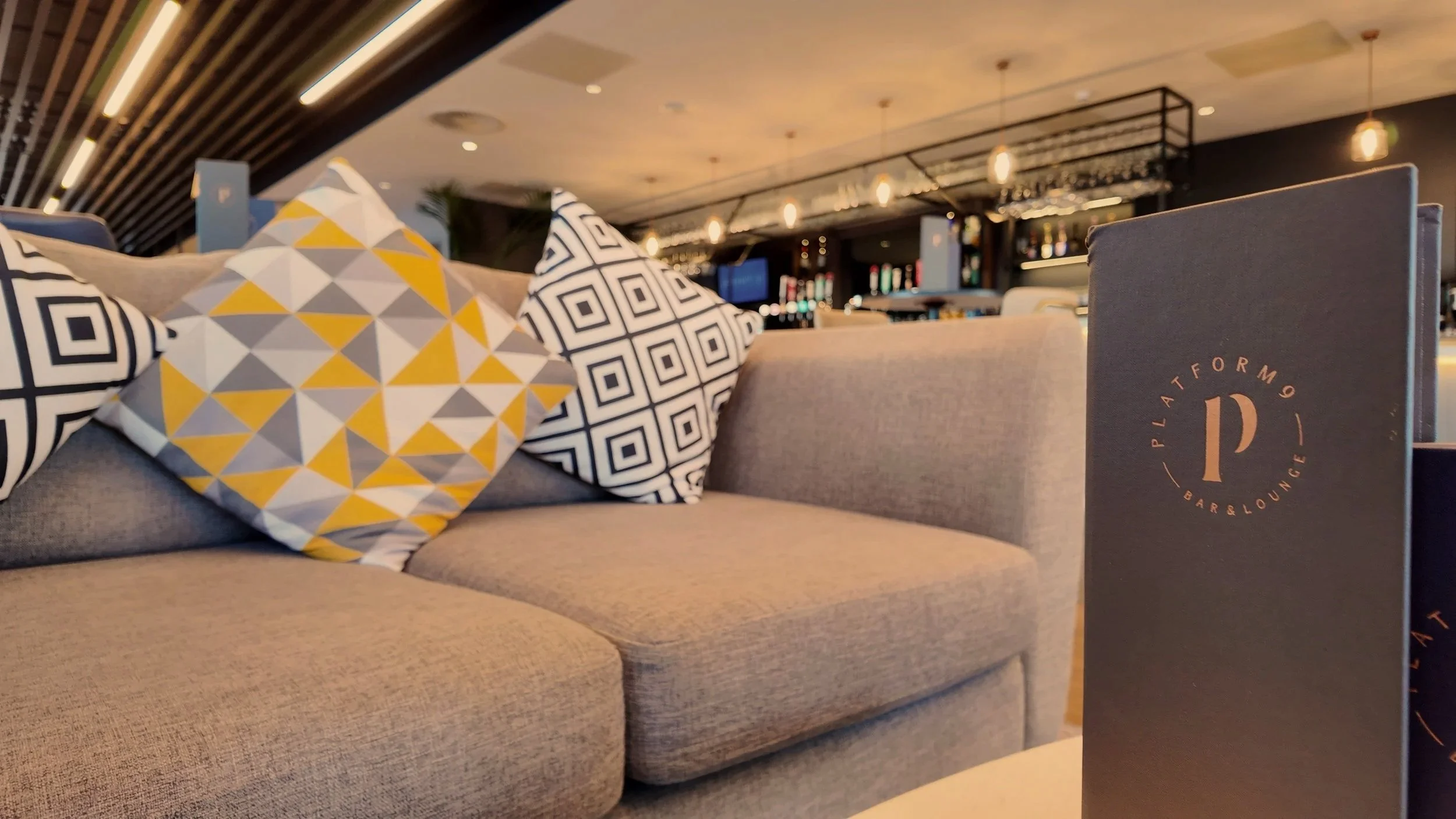A beige sofa with decorative pillows featuring geometric patterns in black, white, gray, and yellow. In front of the sofa, there is a black menu or brochure with a circular logo that reads 'PLATFORM Bar & Lounge'. The background shows a modern bar area with a black ceiling featuring hanging lights, shelves with bottles, and a seating area.