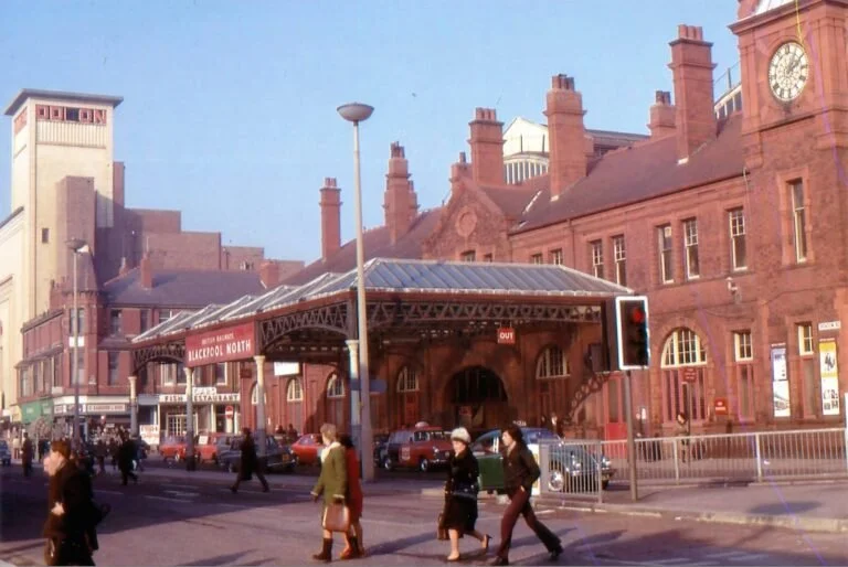 People walking in front of a red brick train station with a clock tower in the background, cars parked along the street, and a traffic light showing red.