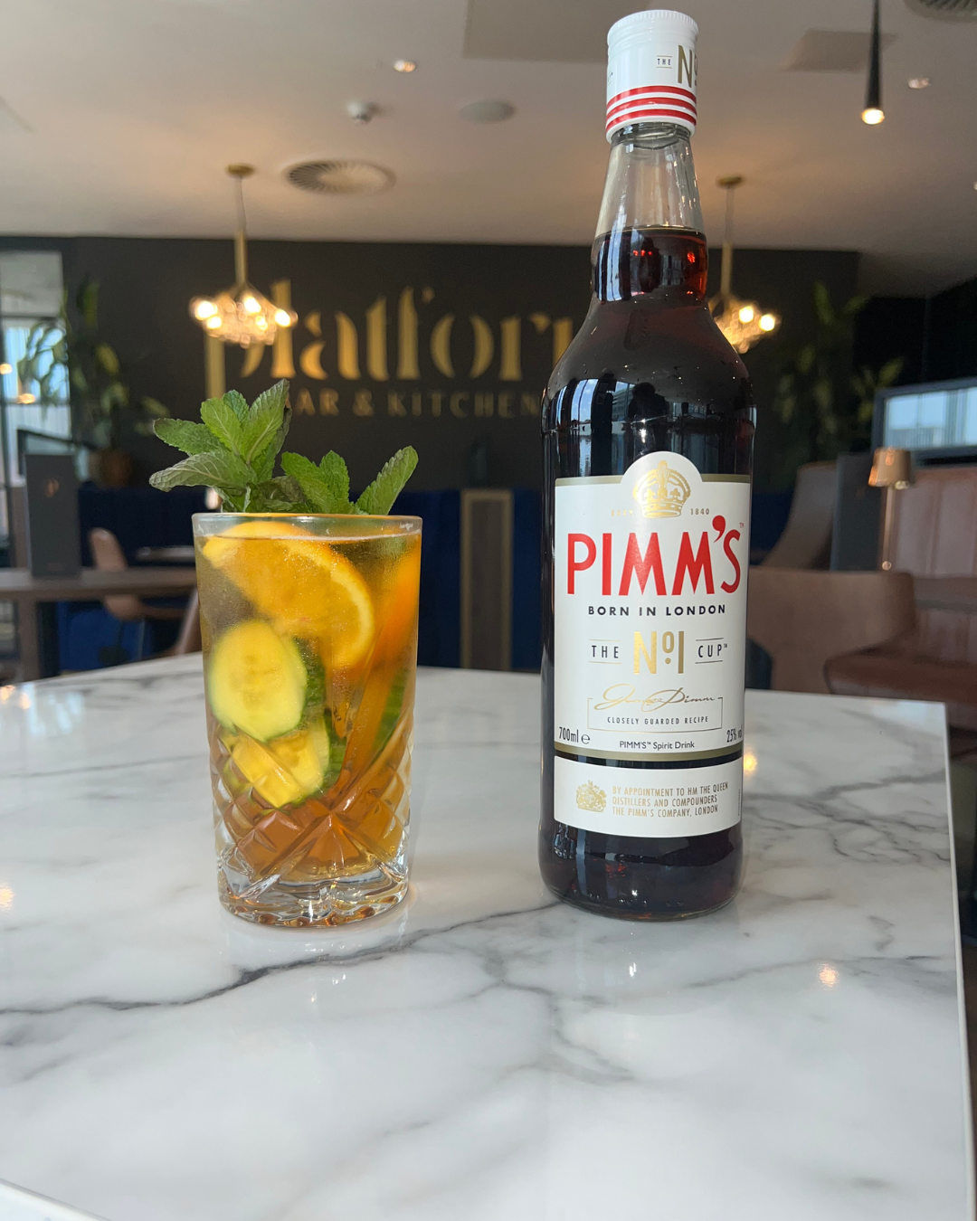 A glass of cocktail with lemon, cucumber, mint, and orange slices next to a Pimm's bottle on a white marble table in a restaurant setting.