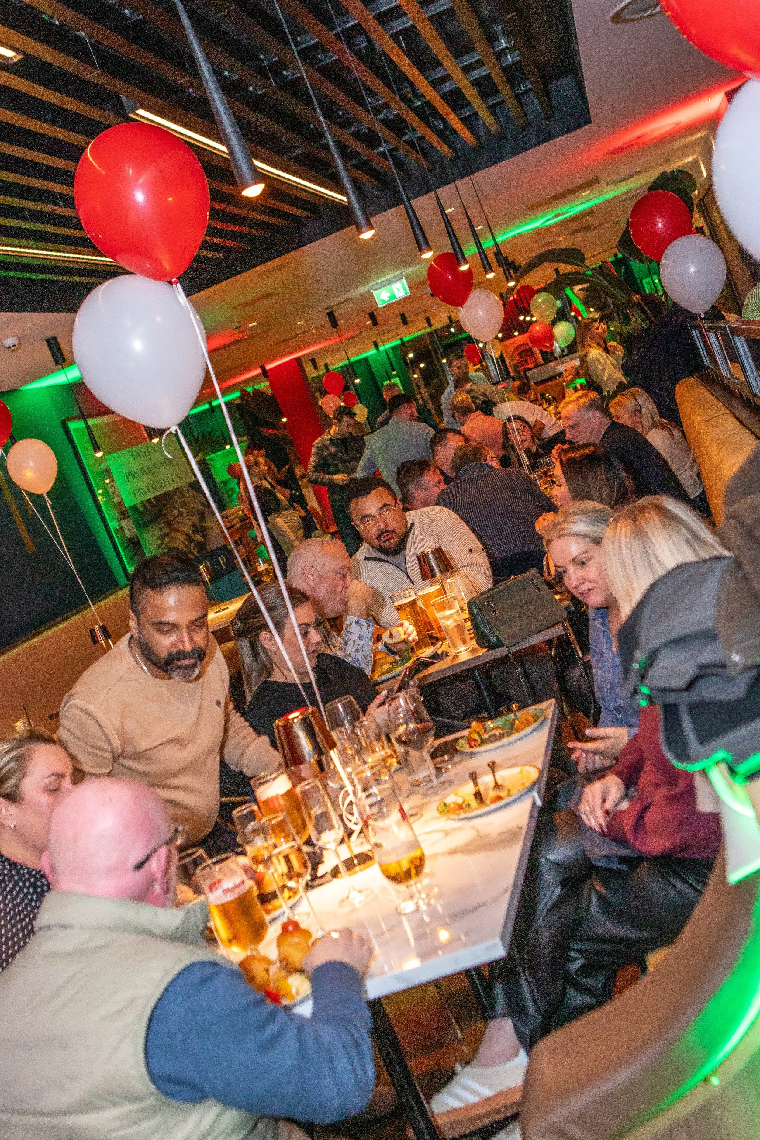A lively restaurant or bar filled with people socializing and enjoying food and drinks. Decorated with red and white balloons, with modern lighting and colorful neon lights.