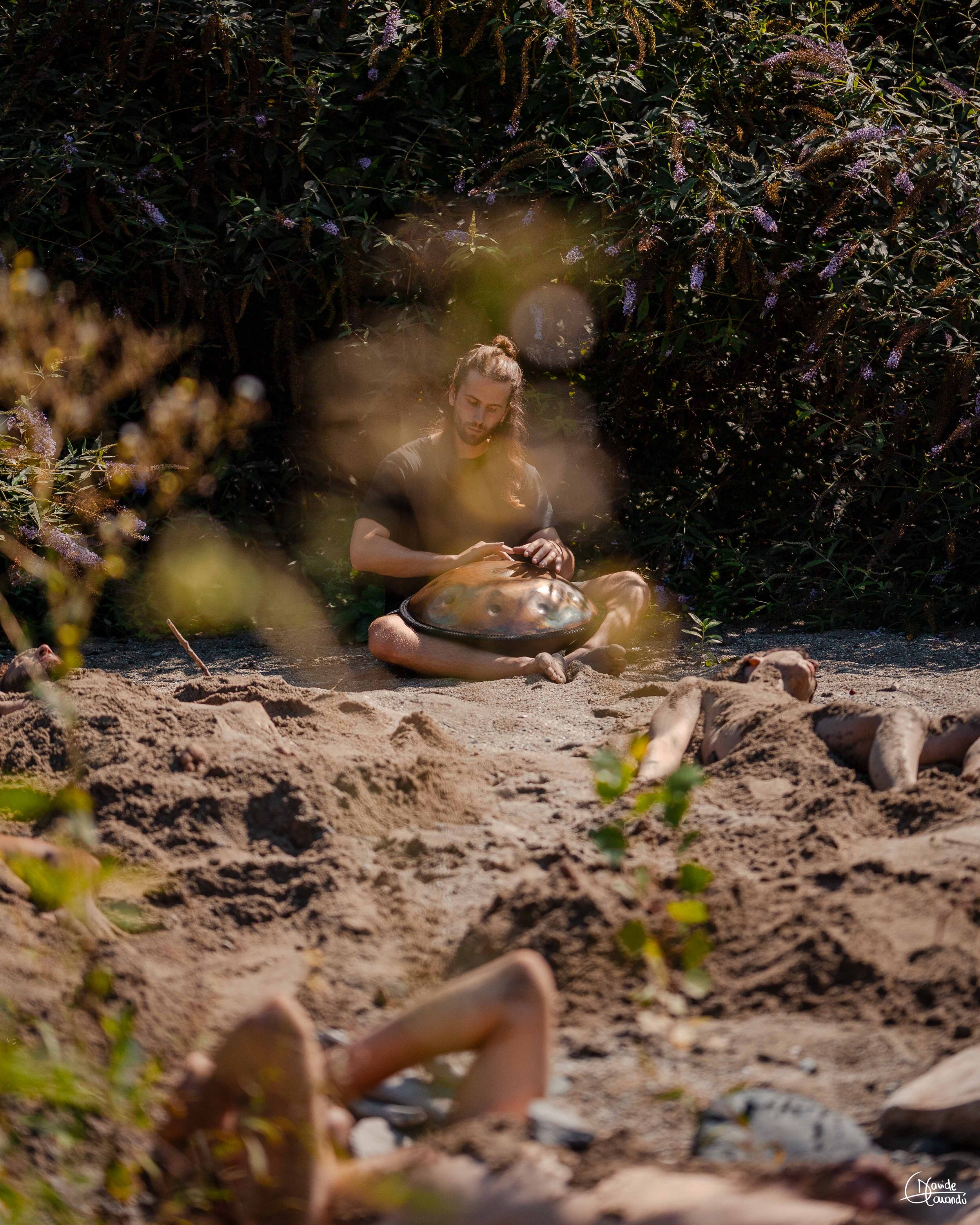 Persona seduta sulla spiaggia con un grande guscio, circondata da vegetazione e sabbia, con altri corpi distesi sulla sabbia in un ambiente naturale.