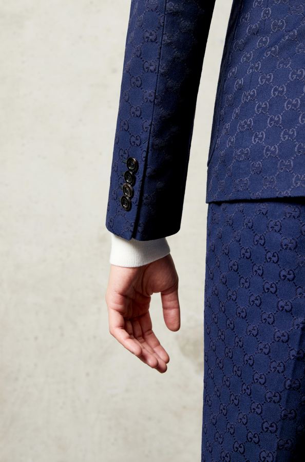 Close-up of a person wearing a dark blue Gucci suit with the signature interlocking G pattern, and a white shirt cuff visible at the wrist.