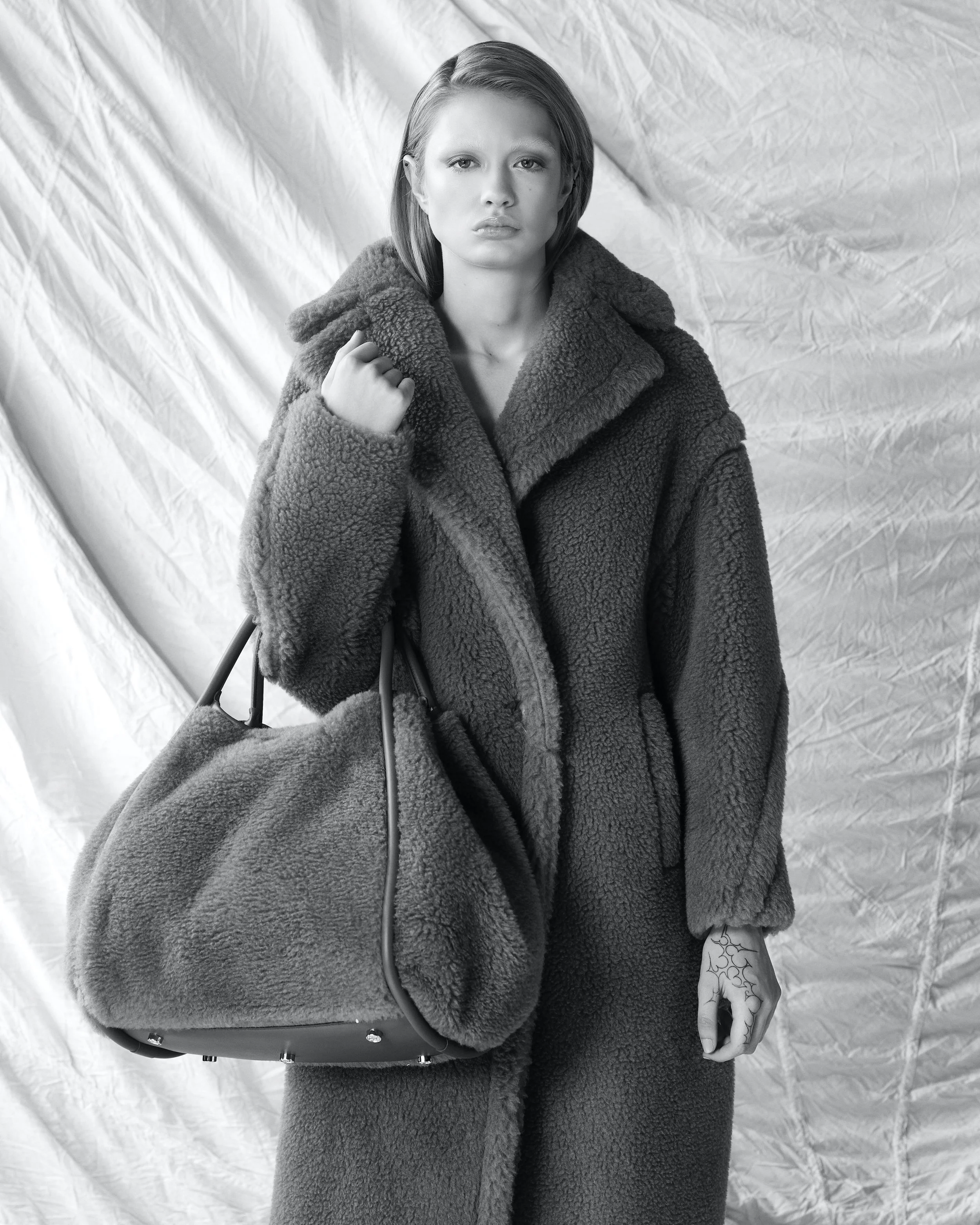 Black and white photo of a woman wearing a thick, textured coat and holding a large furry handbag, standing in front of a crinkled fabric backdrop.