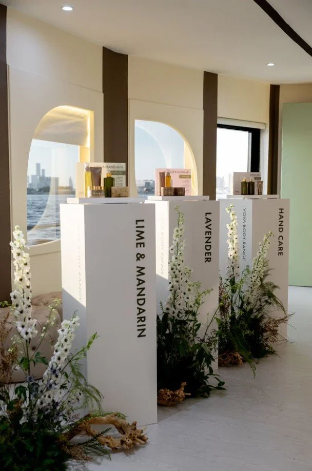 Display stands labeled with different skincare or wellness themes: 'LIME & MANDARIN,' 'LAVENDER,' 'VOYA & BODY RANGE,' and 'HAND CARE,' decorated with white flowers and greenery, set in a bright, modern interior with large windows showing a city view.