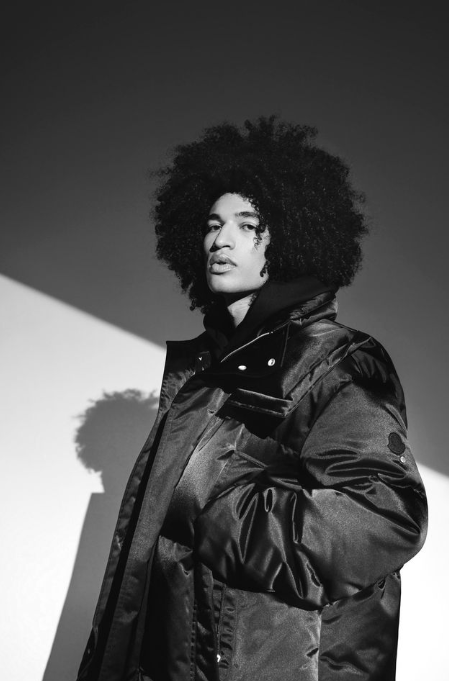 Black and white photo of a woman with curly hair wearing a puffy jacket, standing against a background with strong shadow contrast.