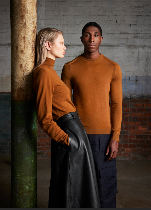 A man and woman standing in front of a brick wall and concrete pillar, both wearing brown tops. The woman is leaning against the pillar with her hands in her pockets, and the man is standing next to her, looking directly at the camera.