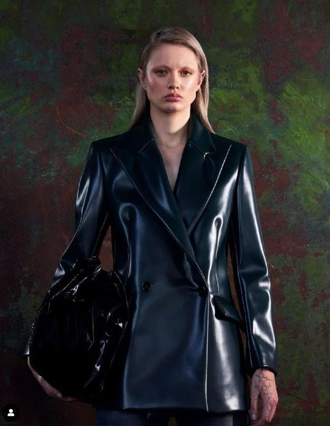A woman with long light brown hair, wearing a black shiny leather blazer, holding a matching black shiny handbag, standing against a dark green and red abstract background.