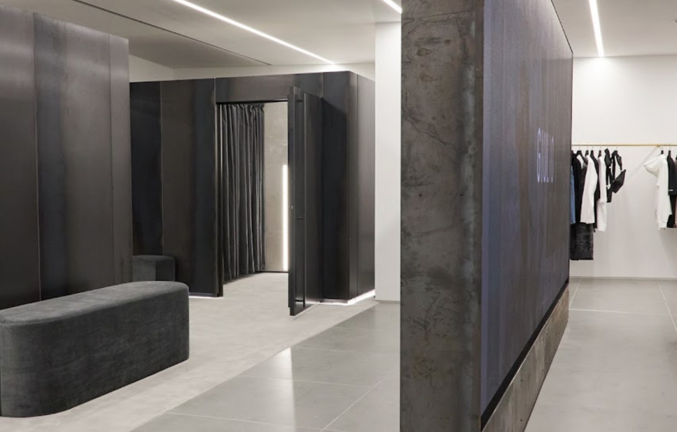 Modern clothing store fitting room area with black curtains, a gray bench, and a rack of clothes in the background.