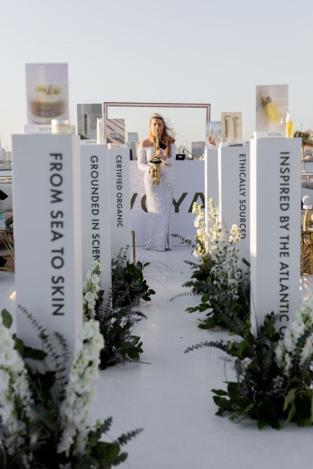 A woman in a white gown playing a saxophone at an outdoor event stage decorated with white flowers and informational banners about the brand VOYA, with a clear sky in the background.