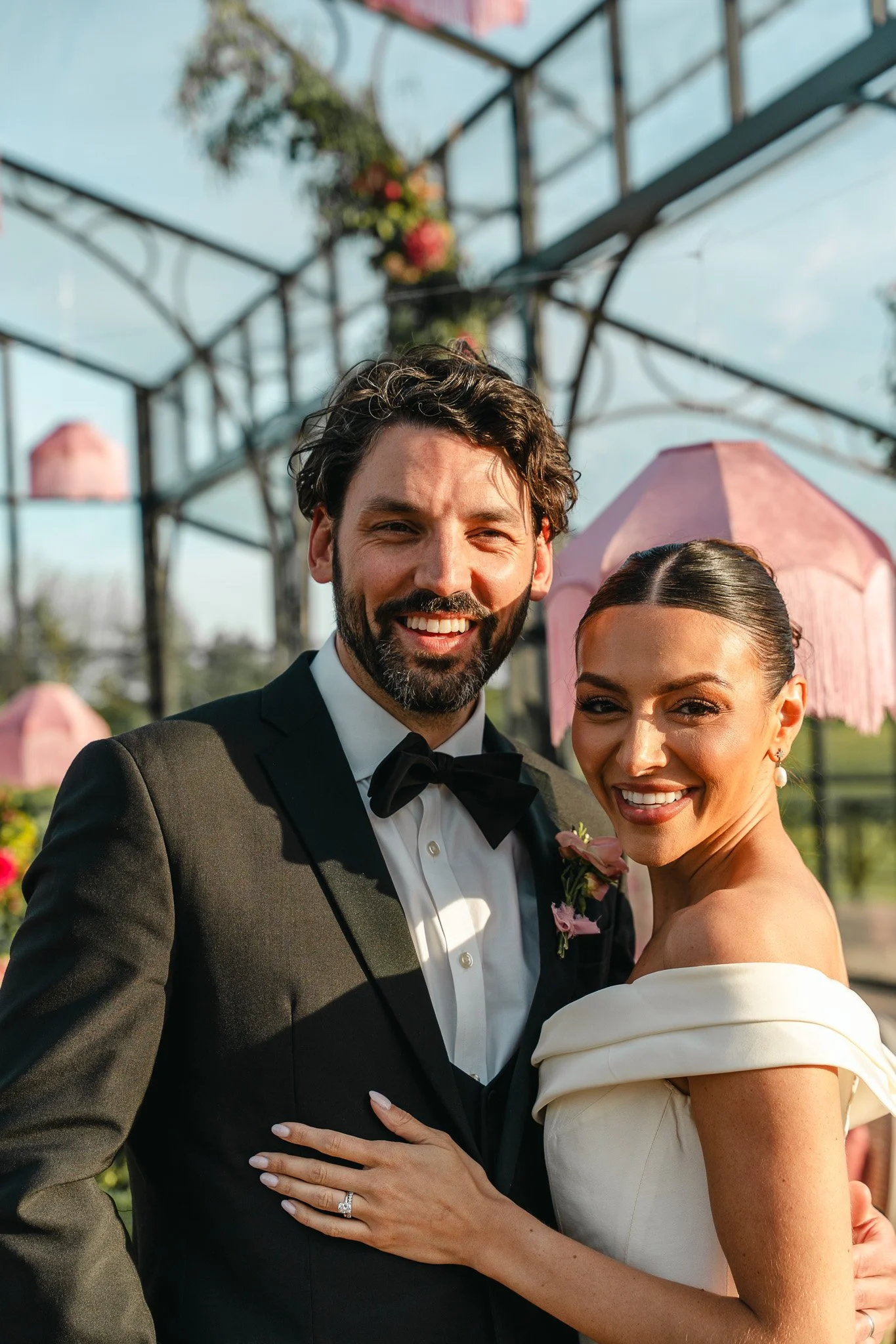 A newlywed couple smiling and posing for a photo outdoors on a sunny day, with industrial-style metal framework and hanging pink lanterns in the background.