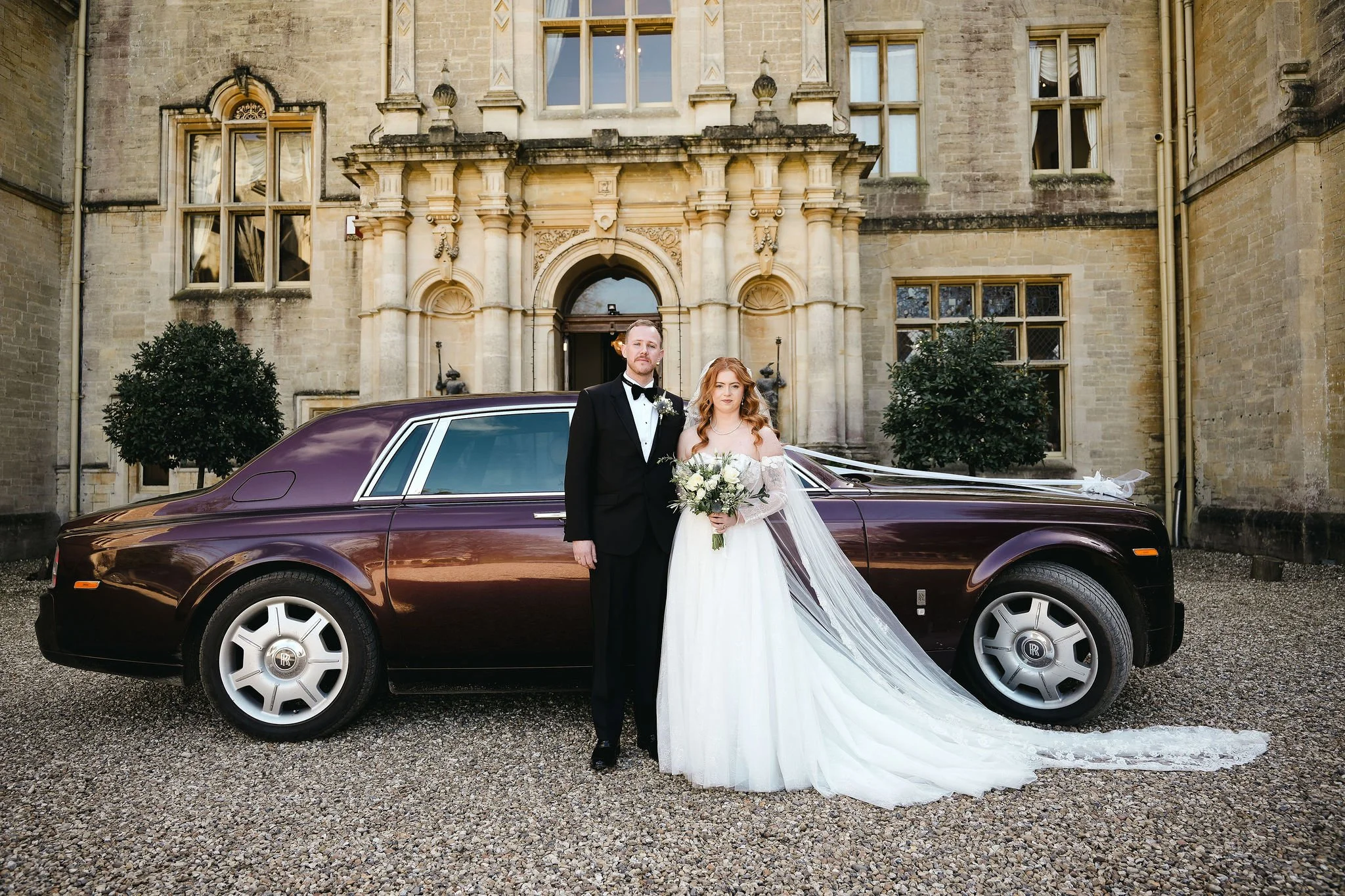 A newlywed couple stands in front of a classic purple Rolls Royce in front of an ornate stone building, with the bride in a white wedding dress holding a bouquet and the groom in a black tuxedo.