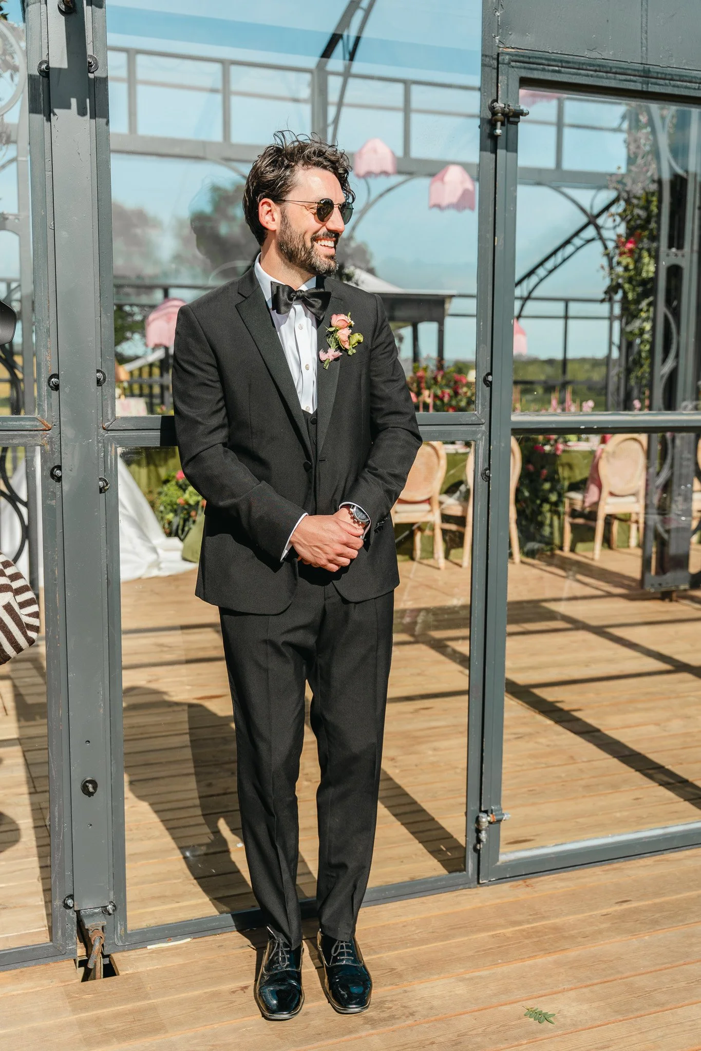 A man dressed in a black tuxedo, white shirt, and bow tie standing outdoors on a wooden deck. He is smiling, wearing sunglasses, and has a boutonniere with pink flowers on his jacket lapel. There are chairs and flowers in the background, and the sky 