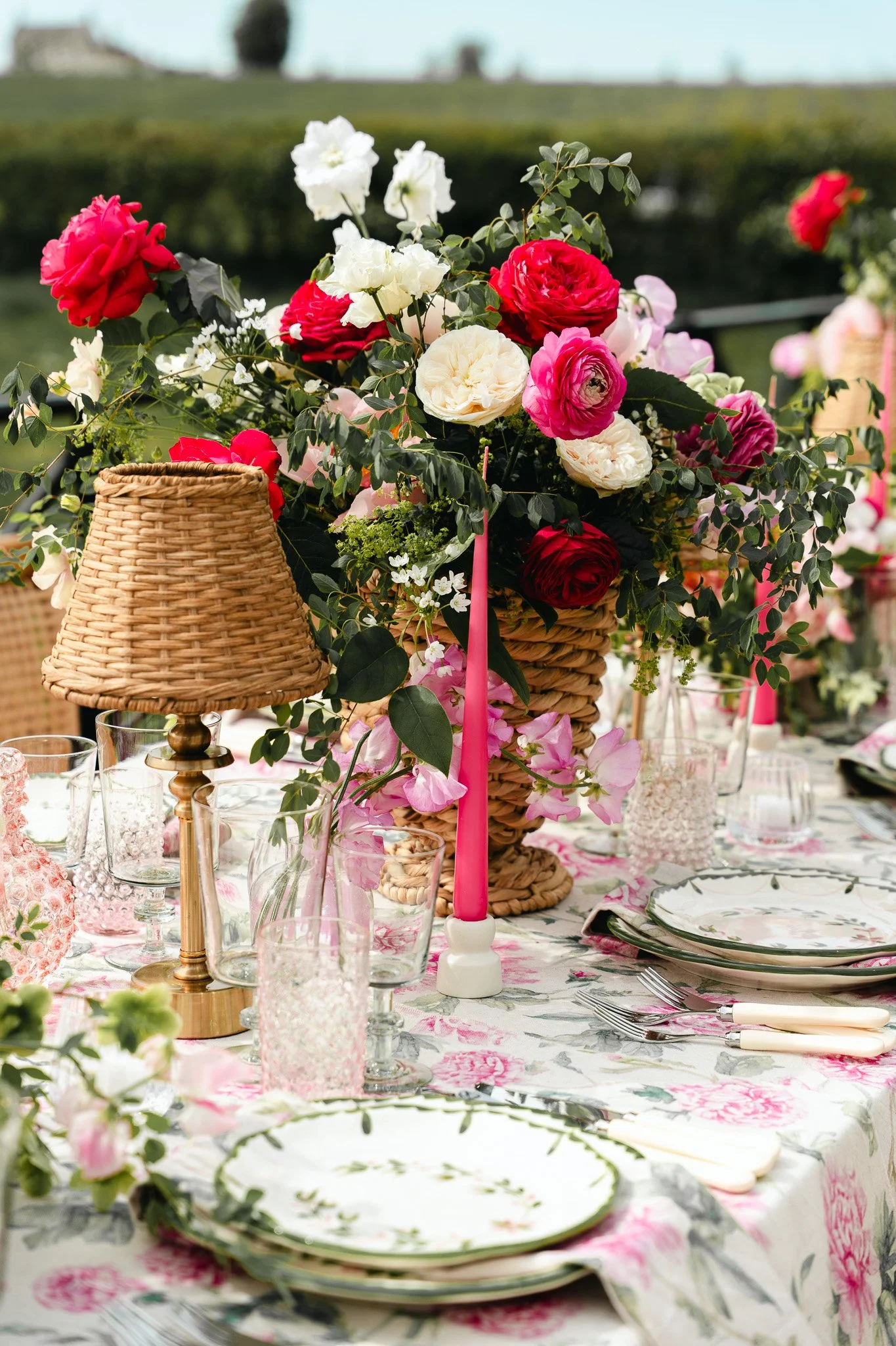 Table setting with floral centerpiece featuring red, white, pink, and purple flowers in a wicker basket, surrounded by pink candles, glassware, and floral-patterned plates and napkins.